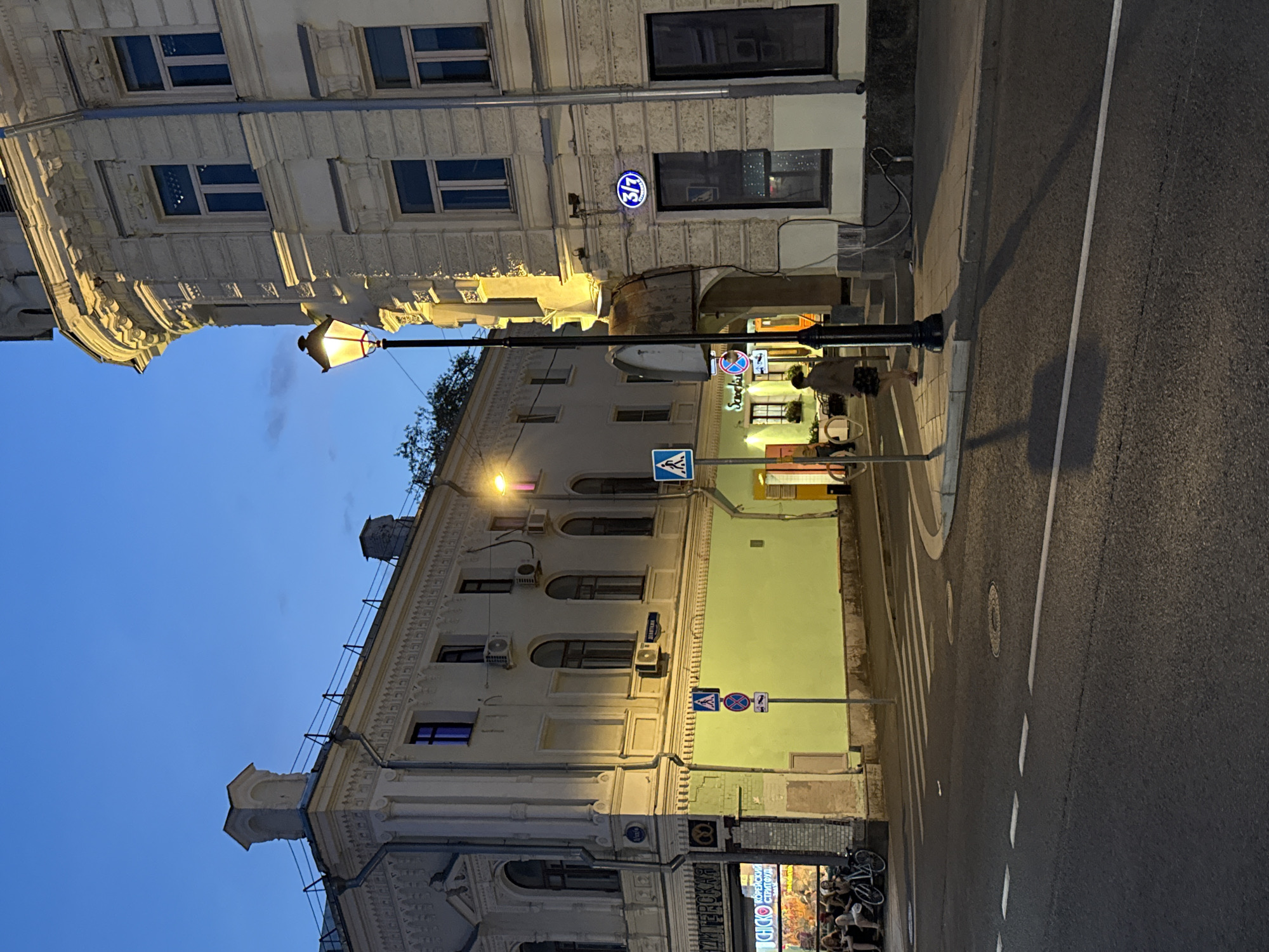 The image depicts a street scene in Moscow, Russia, at dusk. On the right, a grand, light-colored building with ornate architectural details and multiple windows dominates the frame. A vintage-style lamppost stands in front of it, casting a warm glow. Further left, another building, also light-colored, features decorative facades and several windows. A bright green wall is visible on the lower portion of this building.
In the foreground, a street with a pedestrian crossing is visible. Several traffic signs are placed on poles, including a pedestrian crossing sign and a prohibitory sign. On the far left, a group of people are seated outside a cafe or shop with colorful signage. Bicycles are parked nearby.
A lone individual, dressed in a light-colored jacket and a patterned skirt, walks along the sidewalk on the right side of the street. The sky is a deep blue, indicating twilight. The overall impression is a charming urban streetscape in the evening.