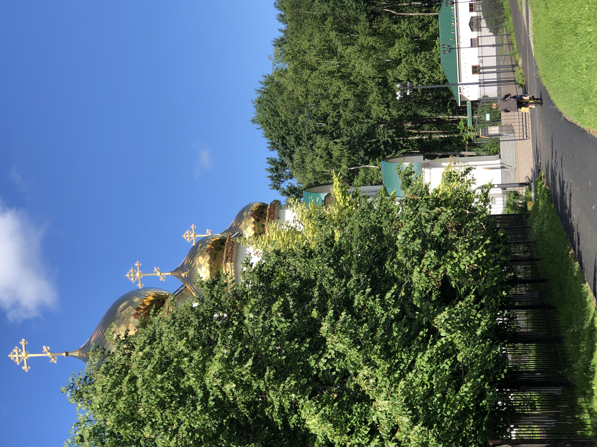 The image shows a view of the domes of the Church of the Resurrection of Christ in Saint Petersburg, Russia, from a nearby park. The church's golden domes and crosses are visible above the lush green trees. In the foreground, a person is riding a bicycle on a paved path. The path leads towards the entrance of the church grounds, which are enclosed by a black metal fence. The sky is clear and blue with a few white clouds. The scene appears to be set on a bright, sunny day.
