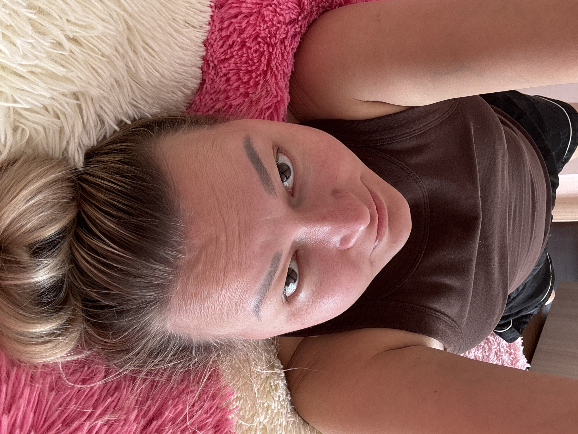 An individual is depicted lying supine on a multi-colored plush surface, consisting of striped sections in vibrant pink and off-white. The person's head is oriented towards the bottom of the frame, with the body extending upwards. The subject is an adult female with fair skin, light brown hair styled in a bun, and she is wearing a dark brown sleeveless top. Her eyes are open, looking directly at the camera. Her arms are raised and partially visible on either side of her torso. Dark shorts or sleepwear are visible above her chest, and a light-colored wall or furniture item is discernible in the upper background. This scene is located in the city of Русский Вожой, Russia.
