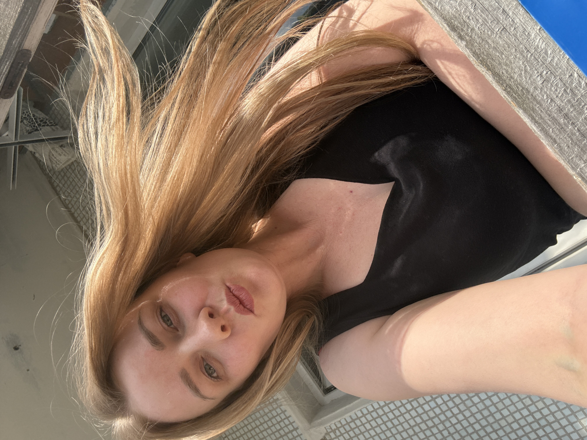 The image displays a person, likely female, with long, light blonde or light brown hair, captured from a high-angle perspective. The photo appears to be a selfie or taken by holding the camera above the subject. Her face is visible in the lower-left quadrant, looking towards the camera with a relatively neutral expression and slightly parted lips. She is wearing a plain black top.
The scene is brightly lit by strong sunlight, which creates distinct shadows and highlights on her hair and skin, indicating daytime. Her long hair, a prominent feature, cascades across much of the image, appearing somewhat windblown or in motion.
The background includes several elements that suggest an outdoor or semi-outdoor setting. In the lower and right parts of the frame, a textured surface resembling white or light-colored tiles is visible, possibly part of a balcony or patio floor. A bright blue object or surface is partially seen on the right, and a metallic or wooden structure, possibly a railing or window frame, is in the upper left background. A reflective surface, which could be water, glass, or a polished floor, is visible near her head and upper left, reflecting parts of the surroundings. The person has fair skin. No text is visible in the image. The overall context points to an outdoor area in Chaikovsky, Russia.