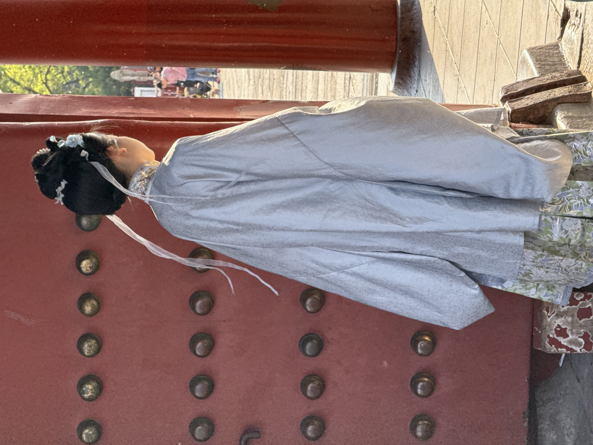 The image shows a woman dressed in traditional Hanfu clothing, seemingly posing or interacting with a structure in the Dongcheng District, China. The structure is red and adorned with rows of metallic studs, with an adjacent element appearing to be stone or concrete, possibly part of a historical building or gate. The background indicates a public area with other people visible, possibly tourists. The woman's clothing is light-colored, with flowing fabric, and she is wearing traditional hair adornments.