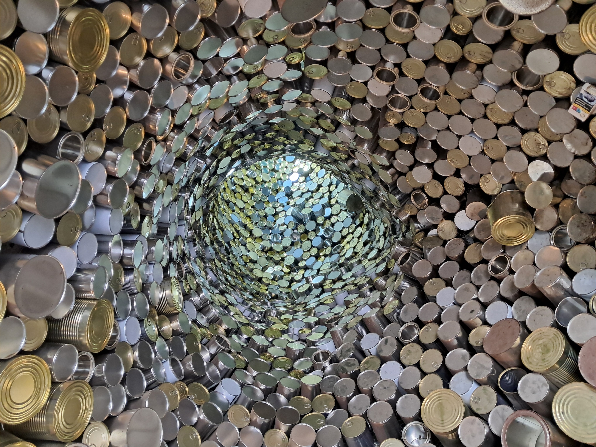 This image depicts an art installation made of stacked and arranged tin cans, forming a circular, tunnel-like structure. The cans are of varying sizes and colors, ranging from silver to gold. The perspective suggests looking into the tunnel, with the density of cans increasing towards the center. The central area exhibits a collection of smaller, possibly differently colored cans, illuminated by a source of light. The installation appears to be indoors, possibly in a public space. This artwork is located in the city of Gabrovo, Bulgaria.