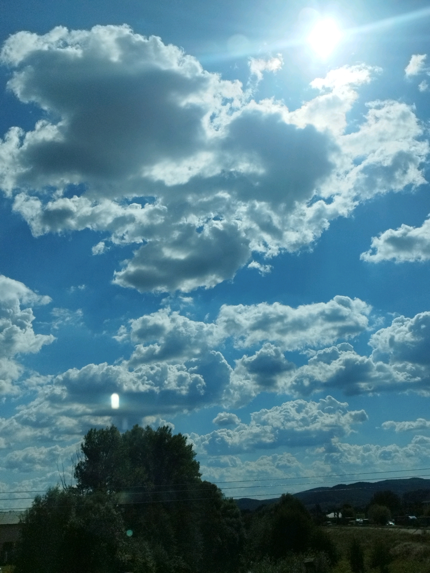 The image depicts a bright, sunny sky filled with numerous fluffy, white clouds against a vibrant blue backdrop. The sun is a dominant feature, positioned in the upper right quadrant, casting a strong glare and lens flare. In the foreground, the tops of dark green trees and some power lines are visible. In the distance, rolling hills are discernible under the expansive sky. The scene appears to be a landscape view, possibly in the outskirts of Curtea de Argeș, Romania, given the natural elements like trees and hills. There are no people or specific actions captured in this image, and the focus is on the atmospheric conditions and natural scenery.