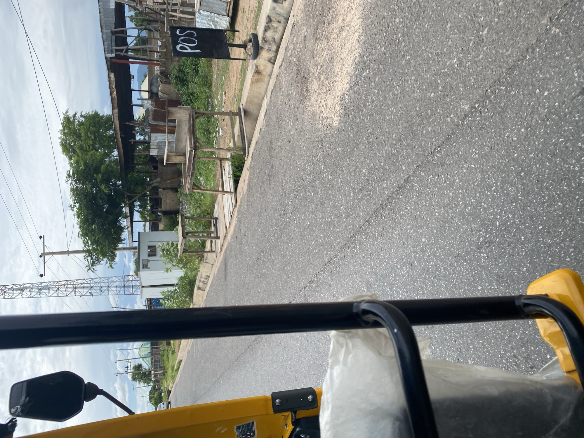 This image depicts a view from a yellow motorized tricycle, likely a "keke-napep" or "Maruwa," traveling on an asphalt road in Jalingo, Nigeria. The foreground shows a portion of the tricycle's yellow body and a black metal frame, with a white bag or tarp inside. The road surface is grey and shows some wear and tear with cracks.
To the right of the road, there is a concrete drainage channel. Beyond the channel, the scene includes a signpost with "POS" written in white chalk on a black background, standing on a makeshift base made from a tire. Adjacent to this are wooden market stalls or tables, some appearing weathered and repurposed. Behind the stalls, there are buildings with corrugated metal roofs and some construction materials. Green vegetation and trees are scattered throughout the area, with a large tree providing shade over one of the structures. In the background, a tall telecommunications tower stands prominently against a cloudy sky. Power lines and poles are visible along the roadside, indicating the presence of infrastructure. A white car is seen parked or driving on the road in the distance to the left. The overall environment suggests a semi-urban or roadside market area in Jalingo, Nigeria, likely during daylight hours.