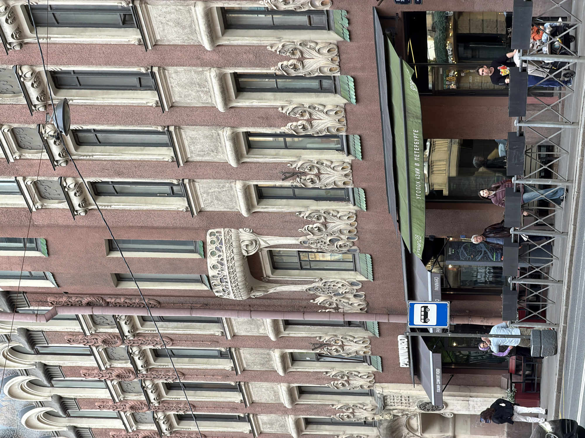 This vertically oriented image captures a bustling street scene in Saint Petersburg, Russia, focusing on the ornate facade of an old, multi-story building. The building is painted in a muted reddish-brown, heavily adorned with intricate, light-colored stucco or stone carvings, particularly around its numerous windows. These decorative elements include elaborate scrolling motifs, classical cornices, and sculptural accents, indicative of historical European architecture.
At street level, the building hosts commercial establishments with outdoor seating areas. A green awning bears the Cyrillic text "УГОЛОК АЗИИ В ПЕТЕРБУРГЕ" (A Corner of Asia in Petersburg), and another sign reads "PANINARO" with "ITALIAN COMFORT FOOD" below it, hinting at diverse culinary offerings. Metal barriers and outdoor tables and chairs are visible on the sidewalk, where several people are present. One man in a black shirt with a red logo is standing by an outdoor table, while others are seated or walking. A blue bus stop sign is prominently featured, near which a person in a purple top stands.
The scene depicts a typical urban daytime setting, with soft, possibly overcast, lighting. Power lines or cables crisscross in front of the building, adding to the urban feel. The overall impression is one of a lively street with historical charm and modern commerce.