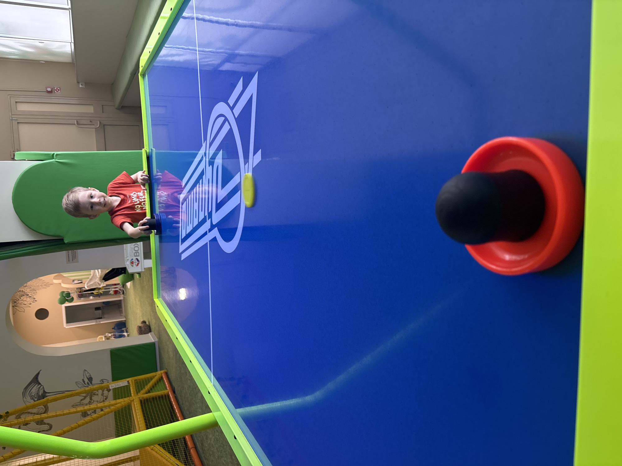 A young boy in an orange shirt stands at an air hockey table with a blue playing surface, playing against an unseen opponent. The table has a lime green border. A puck is visible in the center of the table. The table is located indoors, possibly in a children's play area. The play area also includes green padded surfaces, an arched doorway leading to another room, and a jungle-themed play structure made of yellow and orange tubing. There is an open sign above the arched doorway with various objects inside the room. Location: Chaikovsky, Russia.