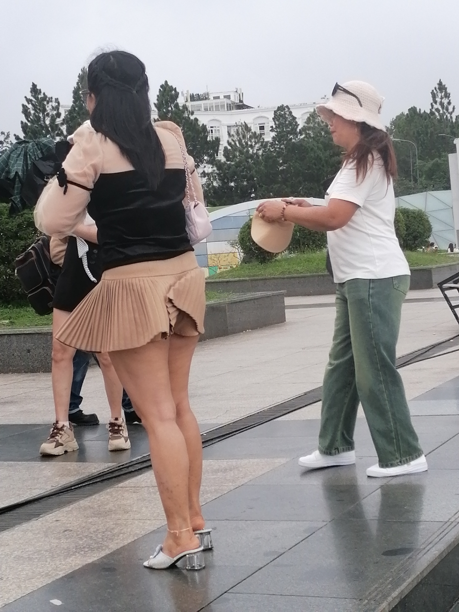 Two women are walking in Da Lat, Vietnam. The woman on the left is wearing a black and sheer-sleeved top with a short pleated tan skirt and white heeled sandals. She has her hair styled in braids and is carrying a pink purse. The woman on the right is wearing a white t-shirt, green jeans, and white sneakers. She has on a light-colored sun hat and is holding a beige hat in her hands. They appear to be walking on a paved pathway with a granite border. In the background, there are trees and a white building with arched windows. The sky is overcast.