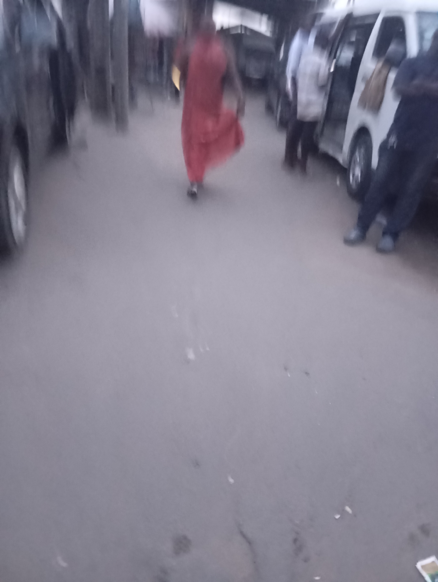 In Port Harcourt, Nigeria, a scene unfolds on a paved street, likely an area with regular vehicular and pedestrian traffic. The image captures a group of people near several parked vehicles, including a white minivan with its door open. The lighting suggests it's either early morning or late afternoon. A person in a vibrant orange garment walks away from the camera, their form slightly blurred by motion. To the right, another individual, dressed in dark clothing, stands with arms crossed, their posture suggesting a watchful presence. The background is indistinct, hinting at the presence of other buildings or structures typical of a city environment. The overall impression is a candid snapshot of daily life on a Nigerian street.