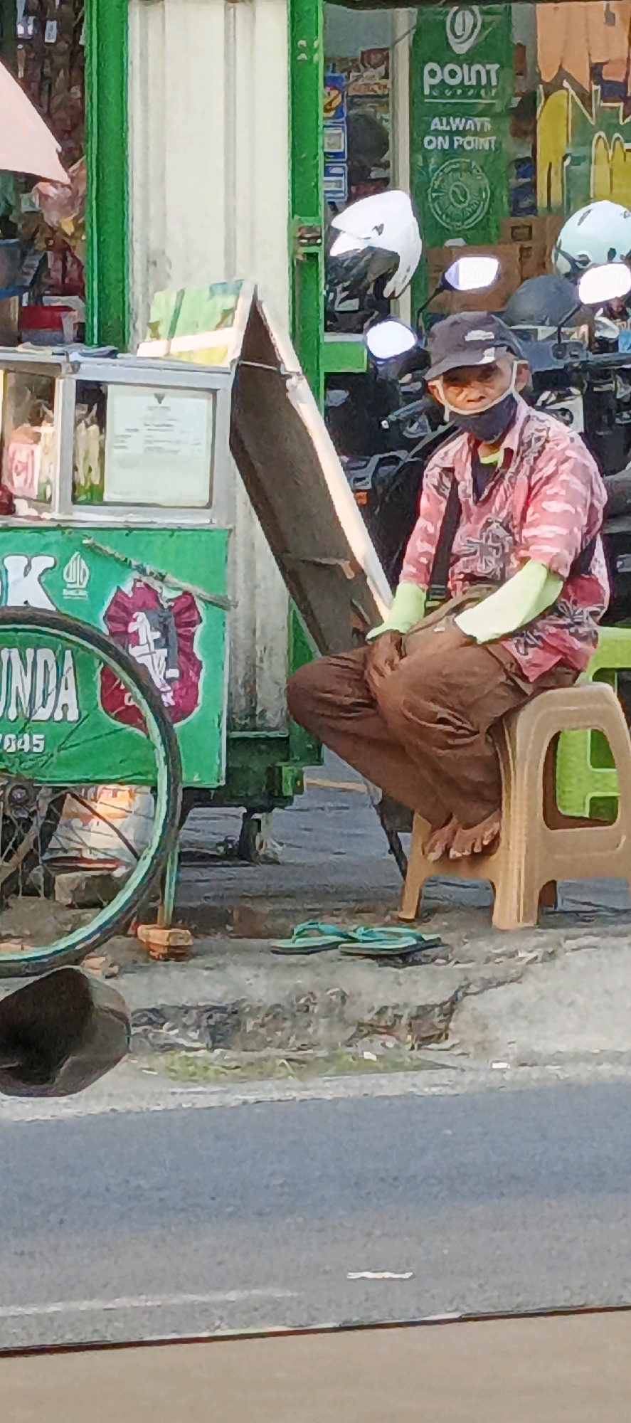 A man wearing a dark baseball cap and a pink patterned shirt with light green sleeves sits on a beige plastic stool in front of a green and white food cart. He is wearing brown pants and a dark face mask. The food cart has a silver-colored display case with items inside and a green wheel with a black tire and rim partially visible on the left. A dark brown object sits on the ground in the lower left corner. A pair of light blue flip-flops lies on the ground in front of the stool. In the background, several motorcycles are parked. A sign with a green logo and the word "POINT" is visible on a white background. The scene appears to be on a sidewalk next to a road. The image is slightly blurry and appears to be taken outdoors during the daytime. This scene is located in Sidoarum, Indonesia.