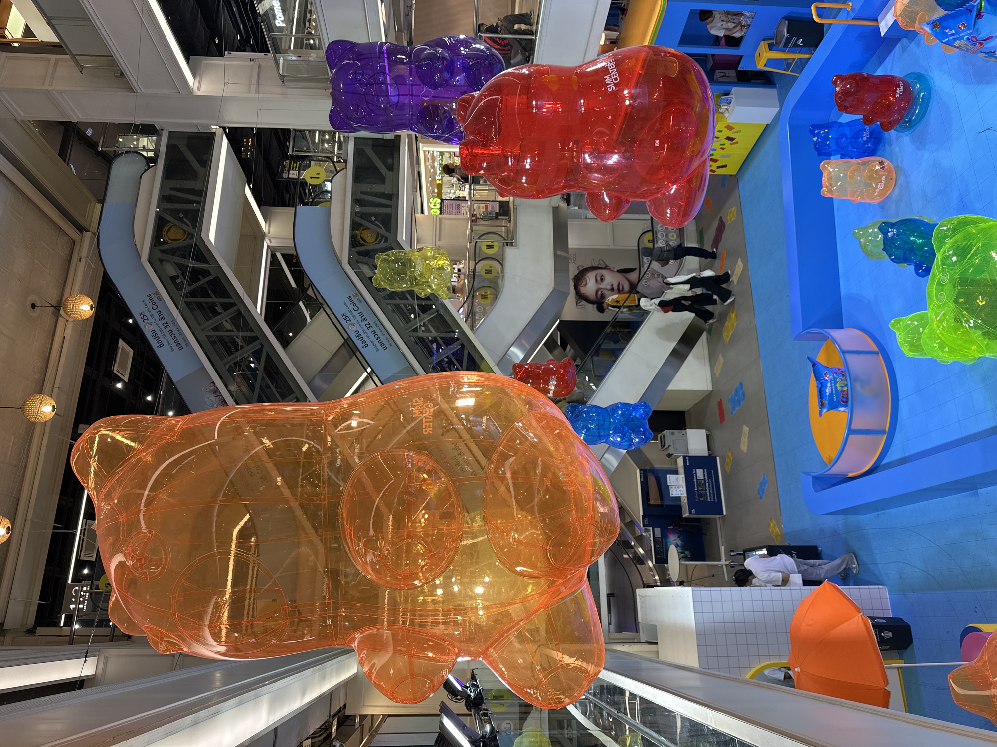 Here is a description of the image:
This photograph captures an interior scene of what appears to be a shopping mall in Bangkok, Thailand. A striking feature is the presence of large, transparent, gummy bear-shaped installations suspended from the ceiling in various colors, including orange, red, purple, and yellow. The mall setting includes multiple escalators, visible through the transparent figures. A woman's face is shown on a large advertisement. Below one of the installations, an umbrella is positioned.
In the background, there is a brightly colored play area, with small plastic teddy bears arranged in a line. A man is lying down on the ground.