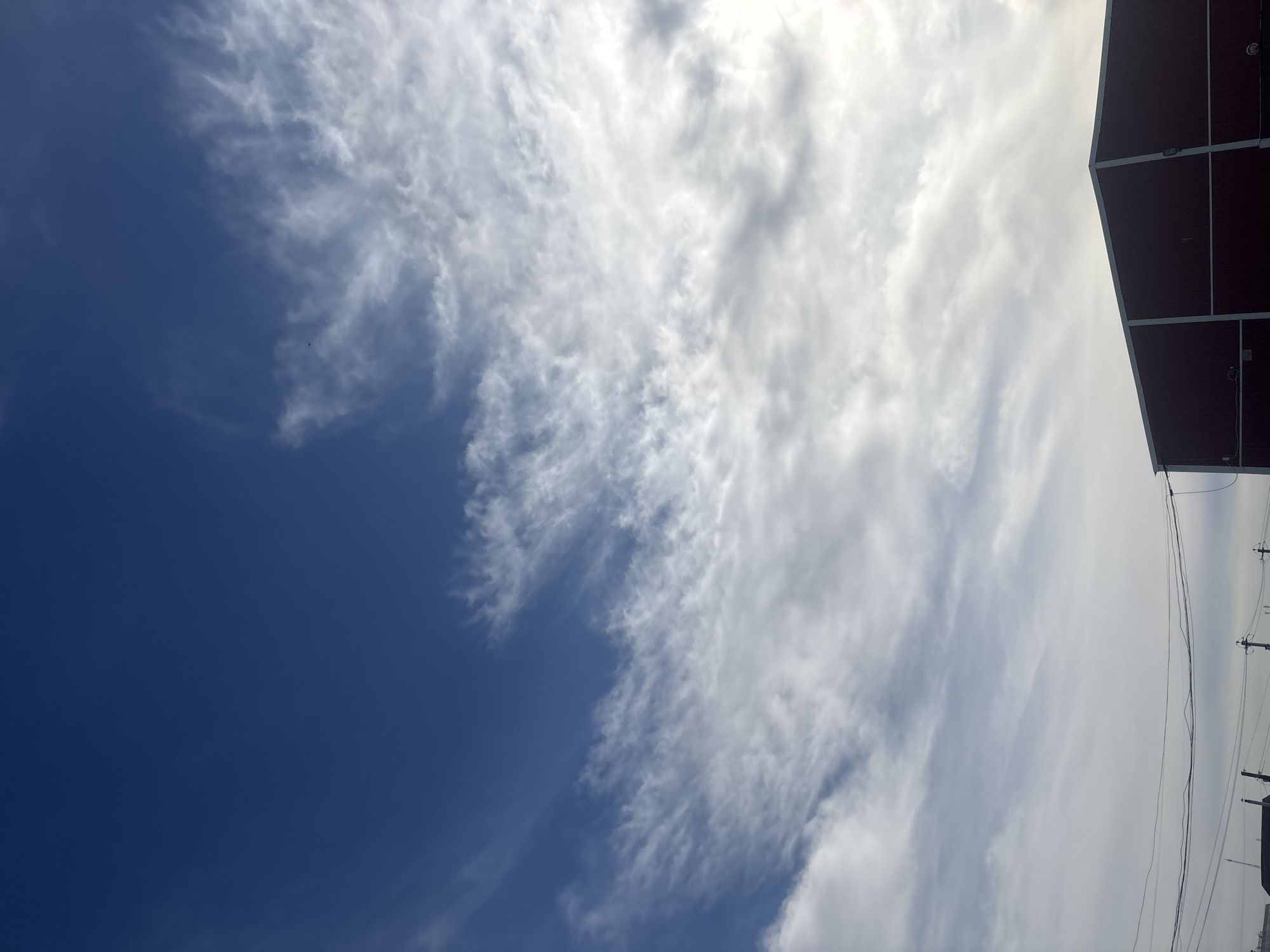 The image shows a sky with white cirrus clouds against a deep blue backdrop. In the upper right corner, a portion of a building, possibly industrial, is visible. Below the building, utility poles and power lines are seen. The image is taken from a low angle, looking upwards. The photograph may have been taken in Chaikovsky, Russia, based on the prompt, but there are no elements within the image to confirm that location.