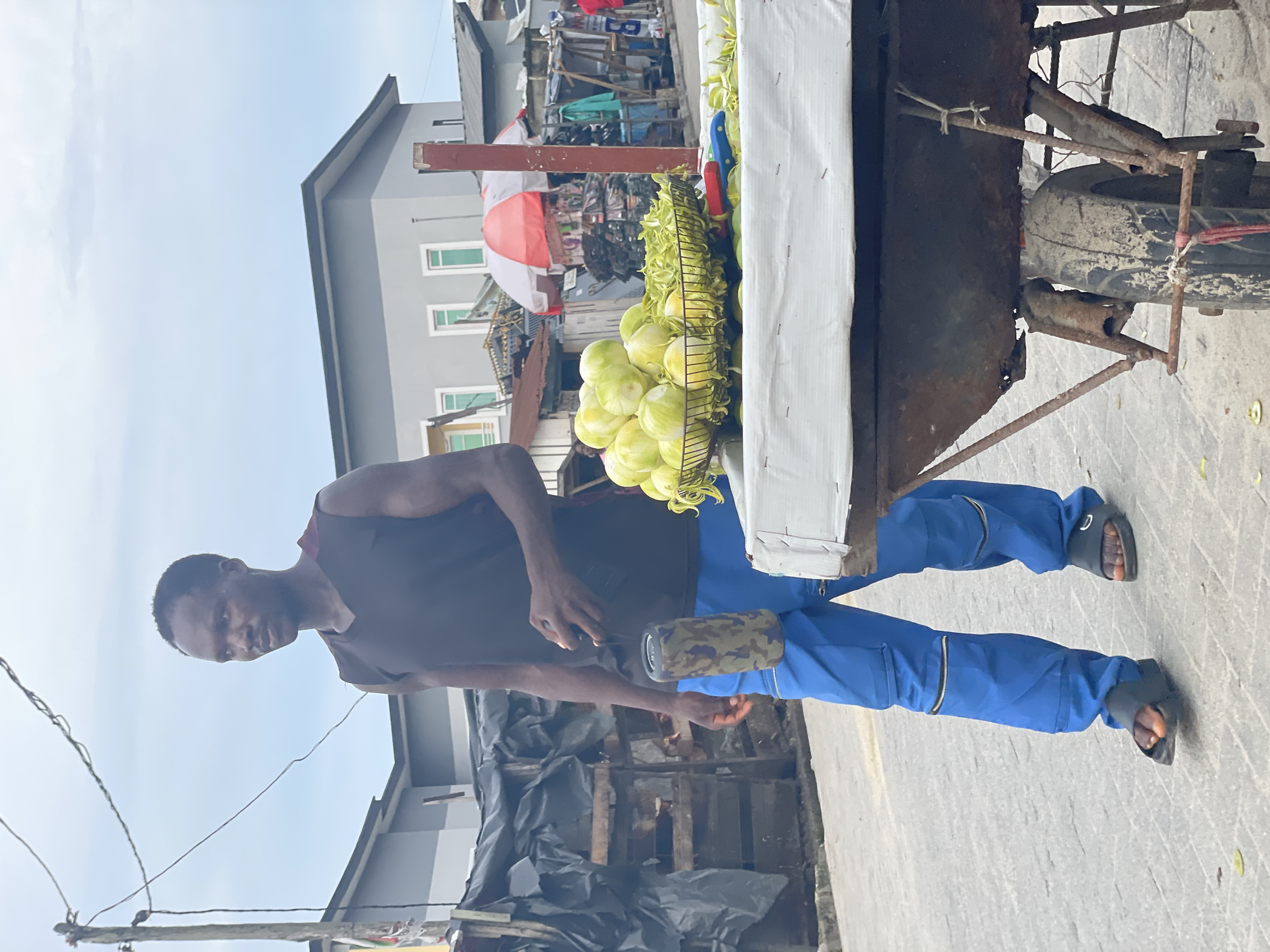 In Ado, Nigeria, a Black man is walking beside a fruit cart. He is wearing a black sleeveless shirt and blue pants with zippers on the legs. He is holding a camouflage-patterned portable speaker. The cart is laden with green fruits, likely tomatoes, and covered with a white tarp. Behind the cart, a red and white umbrella is visible, along with various stalls selling goods. The background shows multi-story buildings with grey and white facades and the cloudy sky of Ado.