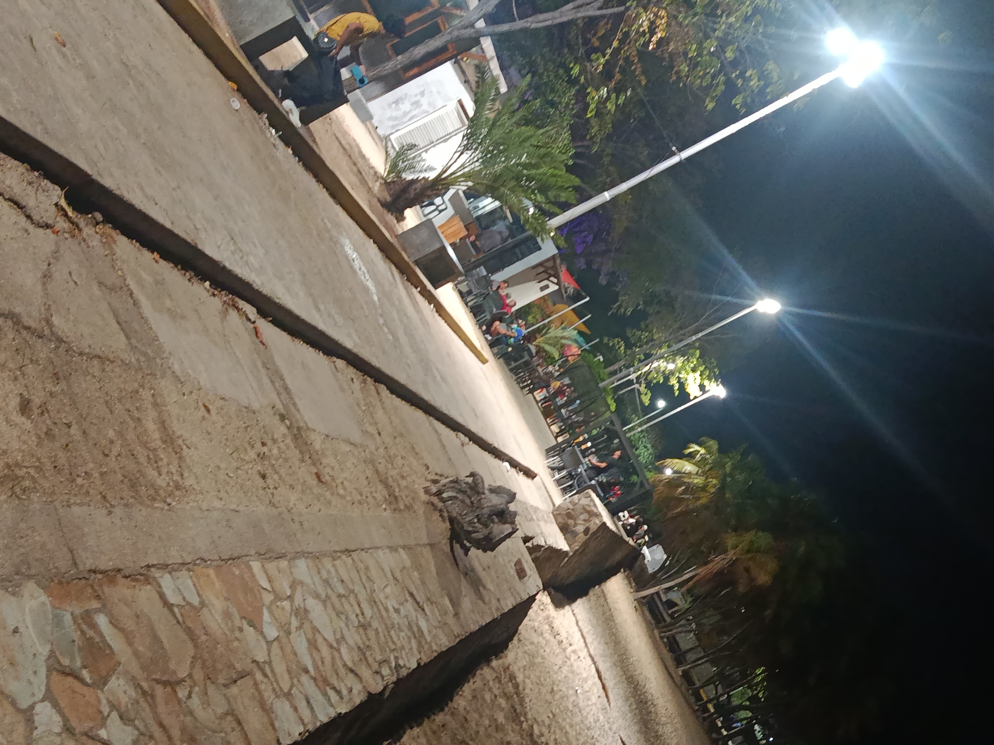 The image depicts a nighttime scene on a waterfront promenade in Juan Griego, Venezuela. The foreground shows a stone and concrete walkway with a deep drainage channel running alongside. Further into the scene, there are several outdoor dining areas with tables and chairs, illuminated by streetlights. People are visible seated at some of the tables. Palm trees and other vegetation are scattered throughout the area. The background is dark, but the sky is illuminated by the bright lights.