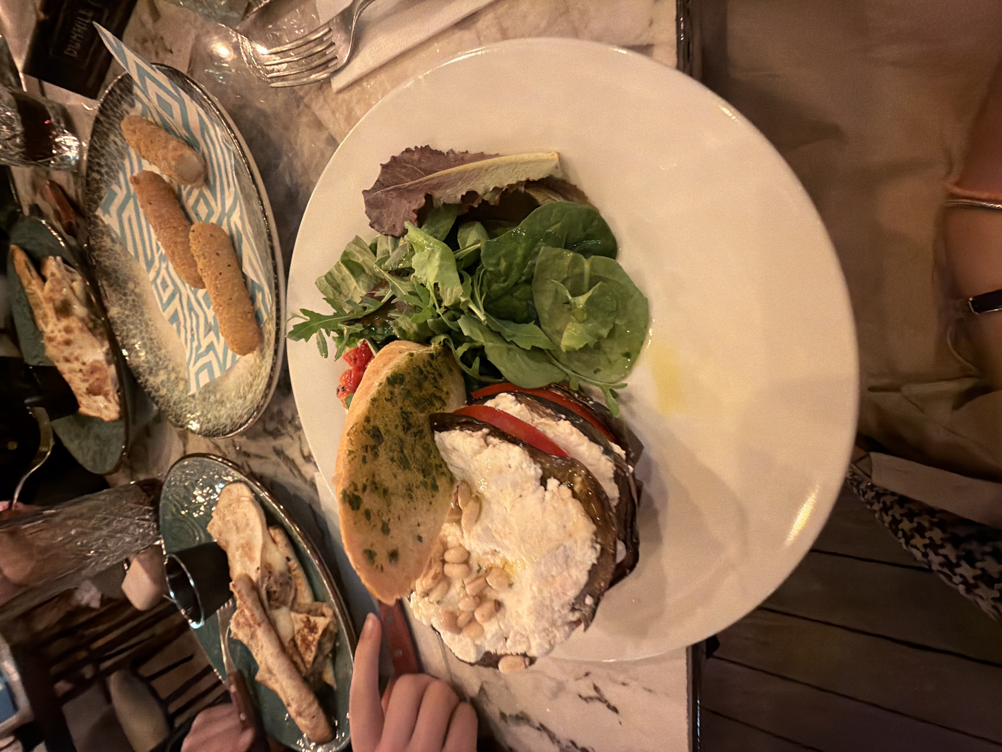 The image depicts a dining table scene, likely in a restaurant. Focus is on a white plate containing a salad with green leafy vegetables, sliced tomatoes, and a stack of eggplant slices with a white creamy topping, possibly ricotta, adorned with pine nuts. Next to the salad is a slice of bread with herbs, possibly garlic bread. Two smaller decorative plates hold breadsticks and pieces of flatbread. A fork and a dark rectangular object, possibly a wallet or case, are visible in the upper left corner. A glass is partially visible. A person's hand is reaching toward the plate, indicating intention to interact with the food. The table appears to be marble-patterned. A fragment of another person's arm and clothing can be seen on the right side.
The specific location in Sofia, Bulgaria is not discernible from the image alone.