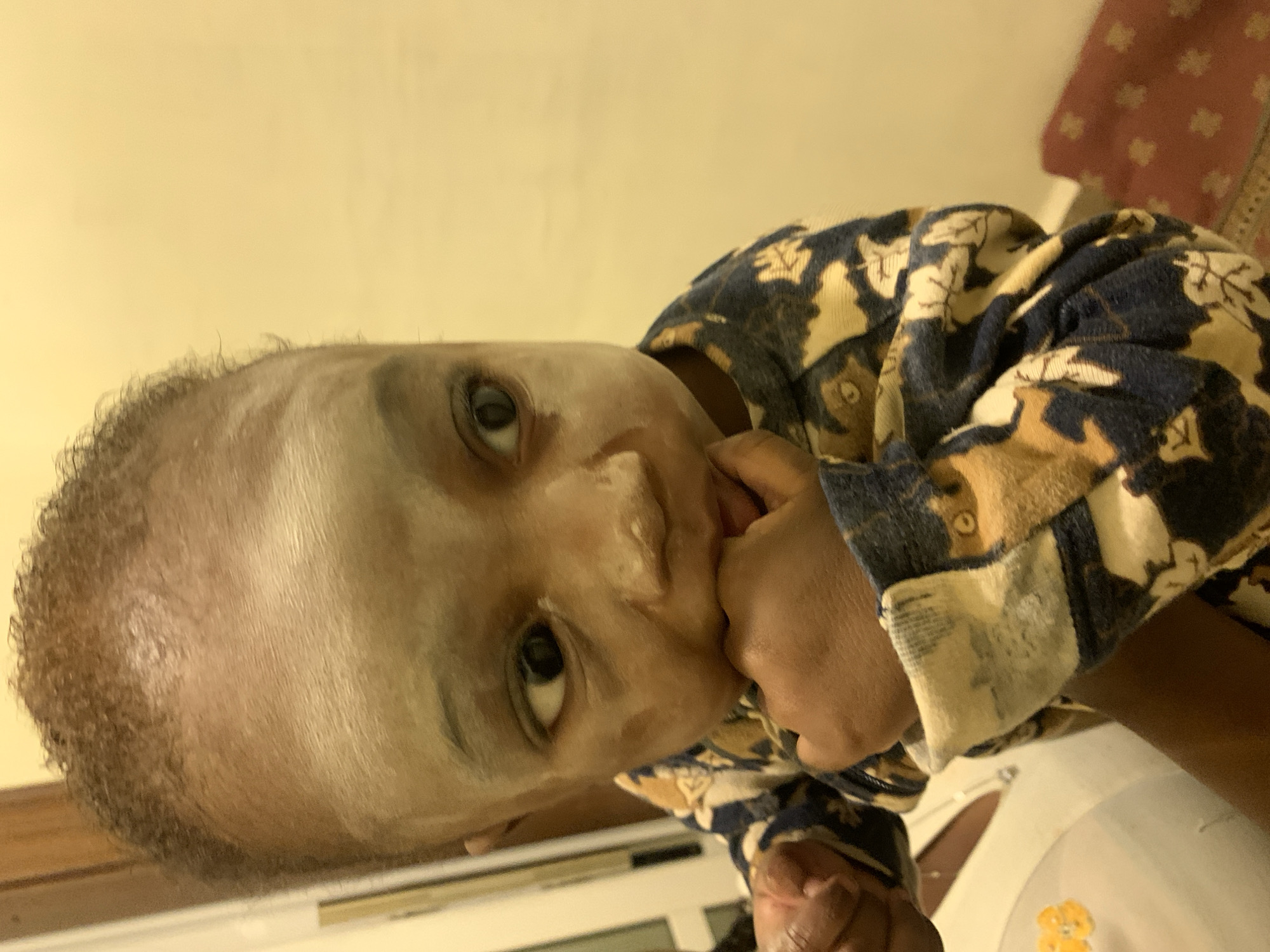 A close-up, rotated image shows a young child with dark curly hair and light-colored cream or paste on their face. The child is wearing a patterned garment featuring blue, beige, and brown colors. The child has their fingers in their mouth. The background shows a beige-colored wall and a hint of a red and white patterned fabric. Beneath the child, a white surface with a small yellow bear sticker is visible. It is not possible to determine the location as Jos, Nigeria, based on the image content alone.