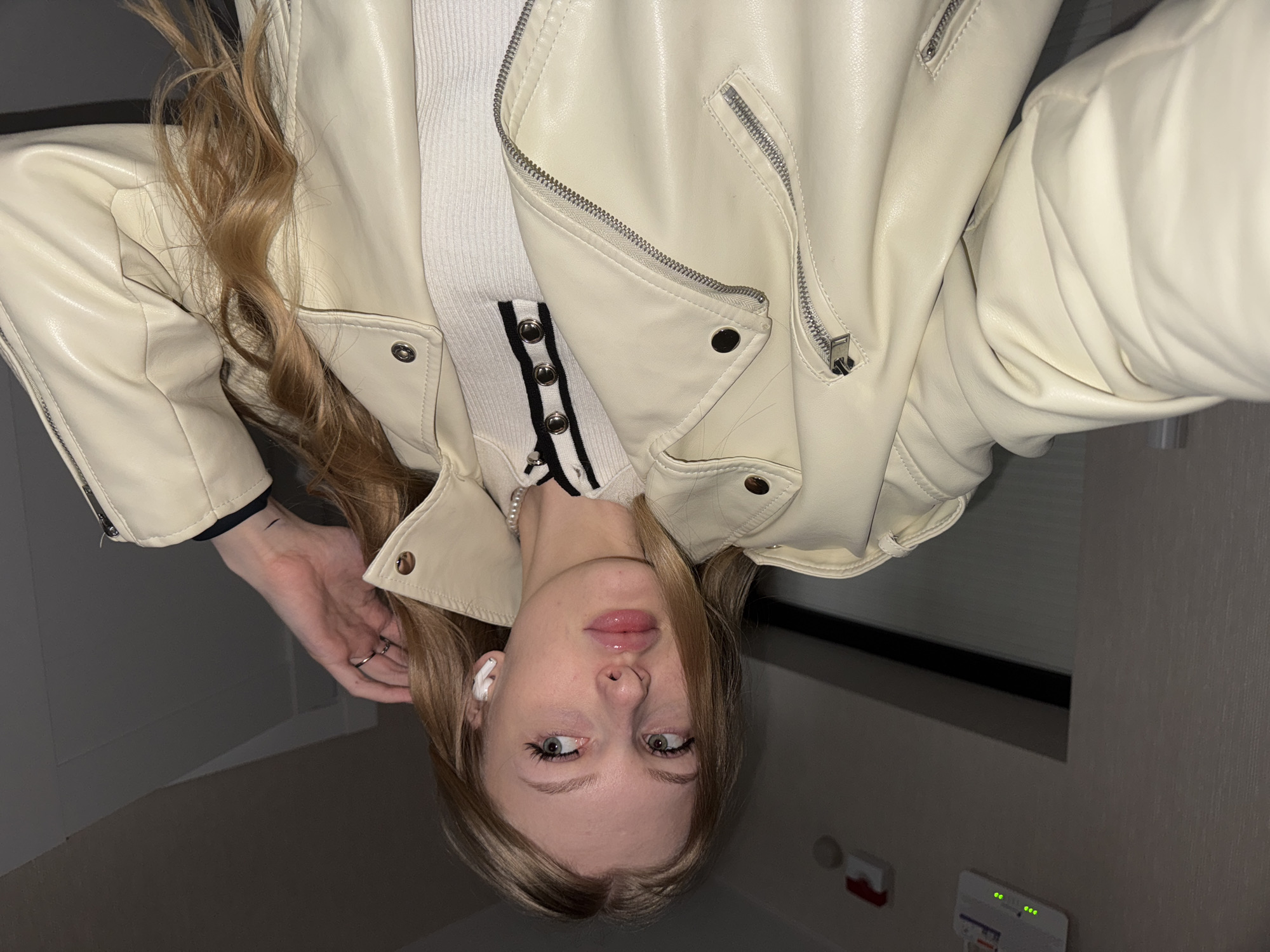 The image depicts a young adult female, located in Perm, Russia, taking a selfie indoors. She has long, wavy blonde or light brown hair and light-colored eyes, gazing off-camera to her right. She is wearing a cream-colored faux-leather motorcycle-style jacket with silver zippers and snap buttons, layered over a white ribbed knit top featuring a black trim and silver buttons along the placket. A single strand pearl necklace is visible around her neck. A white wireless earbud is present in her right ear, and a ring adorns her left hand, which is raised towards her head. The indoor environment features light-colored walls, possibly textured, under subdued lighting. On the upper left wall, a white electronic device with three illuminated green indicator lights is mounted, along with a red wall-mounted object featuring a visible lever.