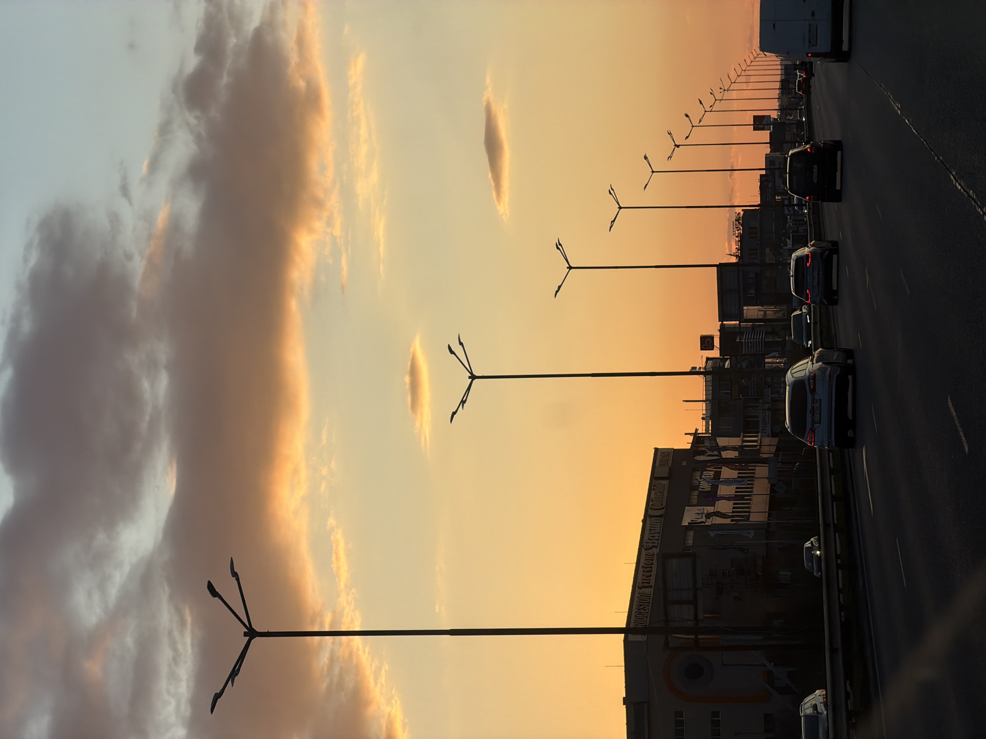 The image depicts a highway scene at sunset or dusk in Sofia, Bulgaria. The sky is a prominent feature, displaying a gradient of warm colors ranging from vibrant orange and yellow near the horizon to softer pale blue and gray higher up. Several clouds are visible; some are brightly illuminated by the low sun, while others appear darker and more voluminous.
Along the left side of the multi-lane highway, a series of streetlights with distinctive multi-arm designs recede into the distance, their dark silhouettes sharply defined against the glowing sky. Cars are present on the road, with several vehicles, including a white van and passenger cars, visible in various lanes, heading away from the viewer. The traffic appears moderate.
To the right of the highway, a line of commercial or industrial buildings extends into the background, largely rendered in silhouette against the warm sky. Visible text on these structures includes "BRIDGES", "FIRESTON", and "FOAM", along with partially visible words like "CONTINENTAL" and "GUMARNA", suggesting businesses related to tires or rubber. The overall atmosphere conveys a sense of transition as day turns into night, blending the natural beauty of the sunset with the functional landscape of urban infrastructure and commerce.