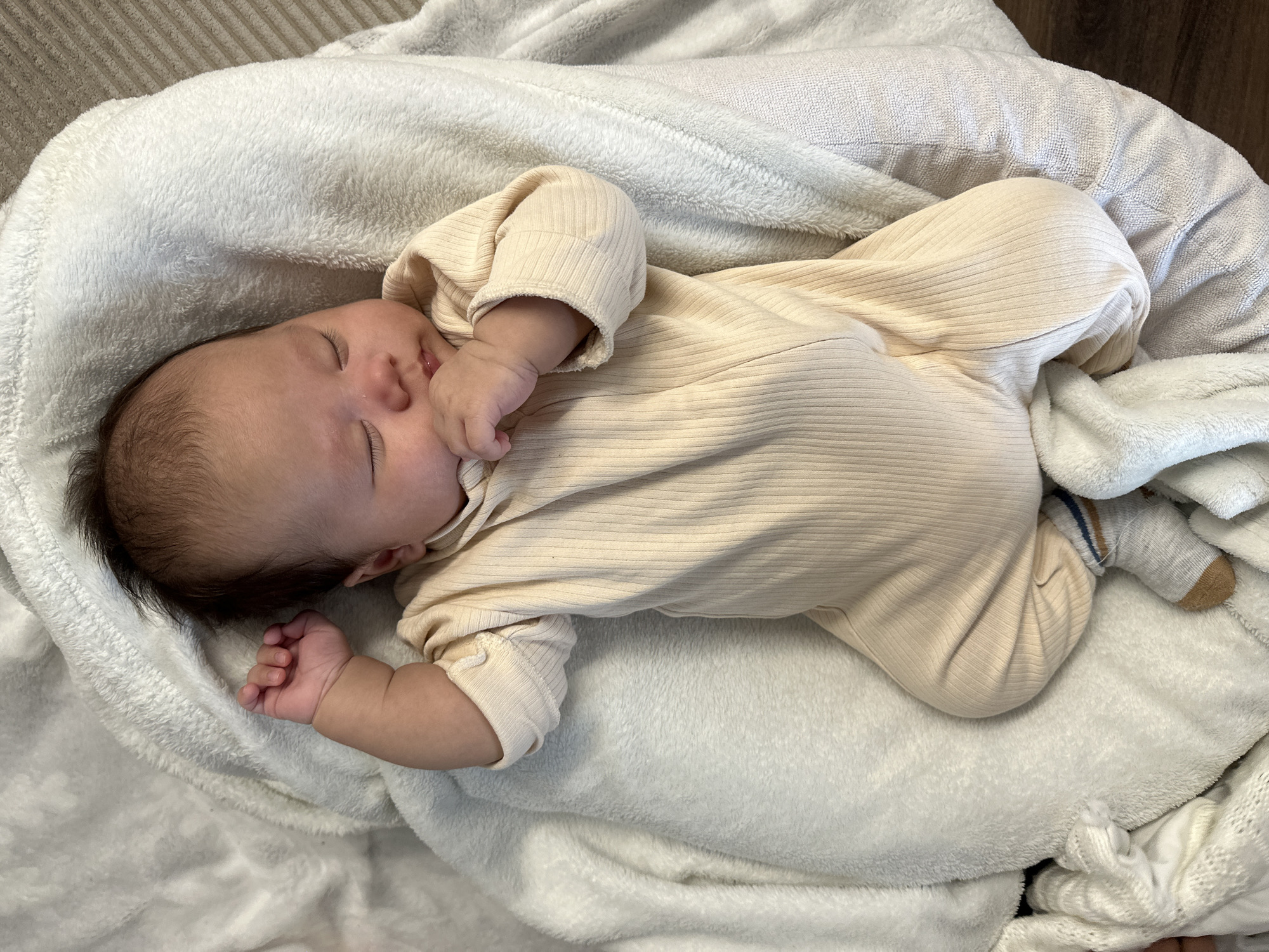 A serene, high-angle shot captures a sleeping baby nestled deeply in soft, white blankets. The baby is lying on its side, with its body gently curled, dressed in a light cream or beige long-sleeved, ribbed bodysuit. One small, chubby hand is raised towards its face, possibly resting near its cheek, while the other hand is clenched into a tiny fist lower down on the blanket. The baby has dark hair and smooth skin, with its eyes closed in peaceful slumber.
The primary blanket enveloping the baby is a plush, fluffy white material, likely fleece or chenille, providing a soft and cozy environment. Hints of other textiles are visible; a ribbed grey surface (possibly a bedspread or sofa) is in the upper left background, and another white blanket with a subtle pattern can be seen towards the bottom right. On one of the baby's feet, a small grey sock with horizontal brown and blue stripes is visible. The lighting is gentle, indicating an indoor setting conducive to rest. The image evokes feelings of tranquility, warmth, and tenderness. There is no visible text.