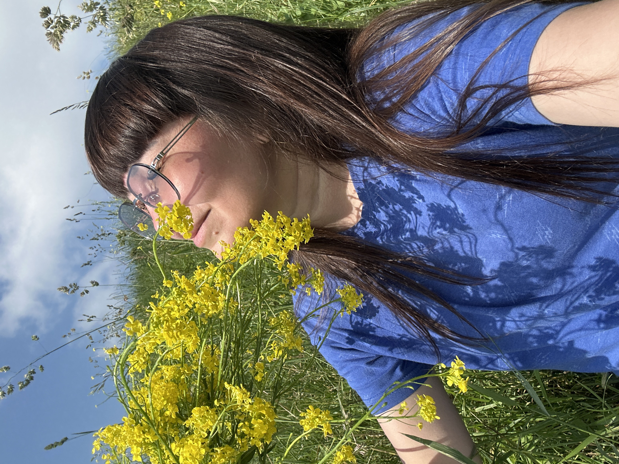 A person with long, dark hair and bangs, wearing a blue t-shirt and pink-tinted glasses, is in a grassy field with yellow flowers. The person is outdoors, with a blue sky and clouds visible in the background. The image appears to be taken in Buranovo, Russia, given the context provided. The person is looking upwards with their eyes closed, seemingly smelling the flowers. The sunlight casts shadows on their face and shirt. The overall scene suggests a peaceful, natural setting.