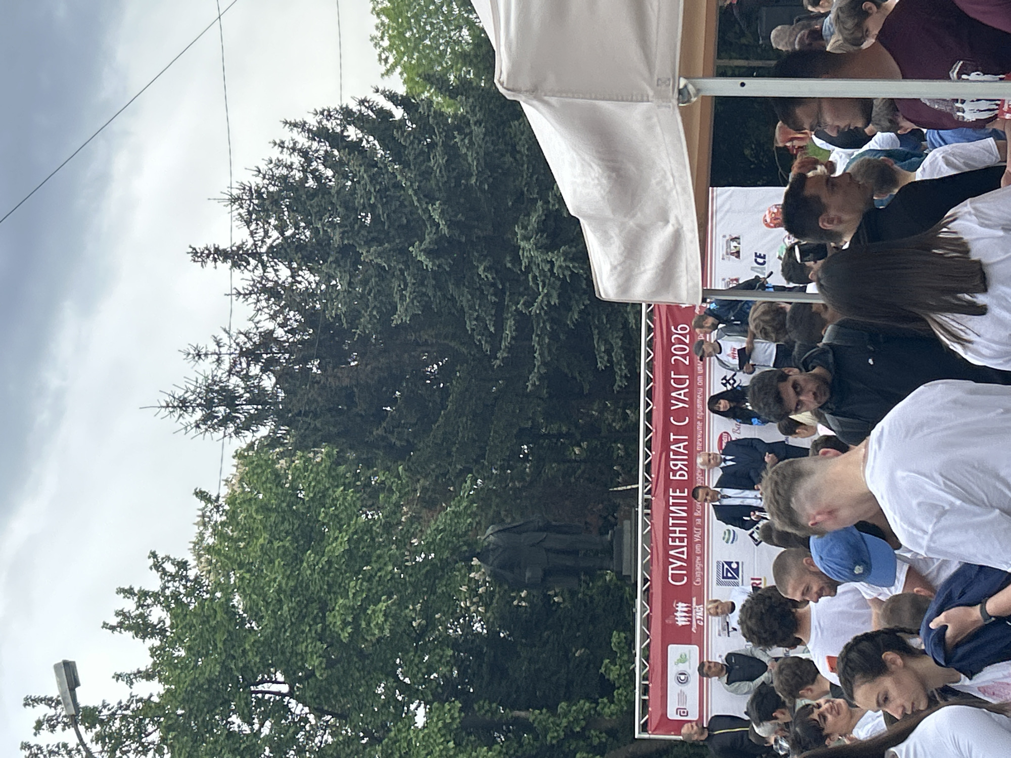 The image depicts a gathering of people, presumably students, in Sofia, Bulgaria. A large banner with the text "СТУДЕНТИТЕ БЯГАТ С УАСГ 2026" ("Students Run with UACG 2026") is visible, indicating a student-related event organized by the University of Architecture, Civil Engineering and Geodesy (UACG). People are seen wearing white shirts and casually dressed. Some individuals are standing around, while others appear to be looking down, possibly at a screen or device. A white tent awning is partially visible in the upper-right corner. Trees are in the background. The statue of a man can be seen amidst the foliage to the left of the banner. The sky is overcast.
