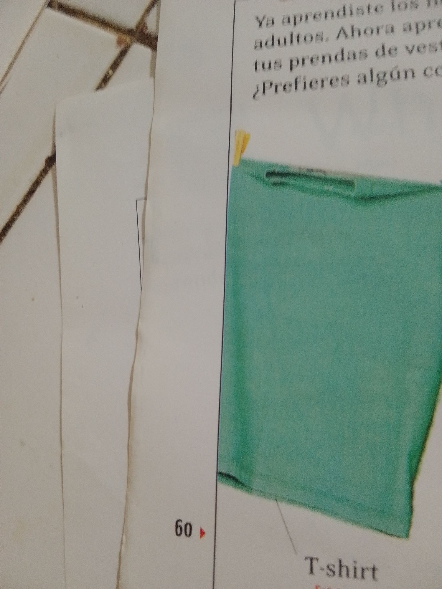 This media file shows a close-up view of a book or magazine page, likely from a children's learning material given the simple text and image. The page features Spanish text and a photograph of a green t-shirt.
The visible text on the page includes: "Ya aprendiste los", "adultos. Ahora apre", "tus prendas de vest", "¿Prefieres algún co", and "60 >>". Below the image of the t-shirt, the word "T-shirt" is also visible, indicating the subject of the photograph. The number "60" followed by an arrow suggests a page number.
The photograph itself focuses on a plain, solid green t-shirt. The garment appears to be a simple design, possibly a plain tee or a part of a larger garment. A small yellow object, resembling a clothespin or clip, is attached near the neckline of the t-shirt, suggesting it might be displayed or held up. The lighting suggests an indoor setting, possibly a classroom or home environment where such educational materials would be used. There are no people or obvious outdoor setting cues visible in this crop. The overall impression is one of an educational resource designed to teach vocabulary or concepts, likely related to clothing. The location context provided, "La Tagua, Venezuela," offers a geographical setting for where this material might originate or be used.