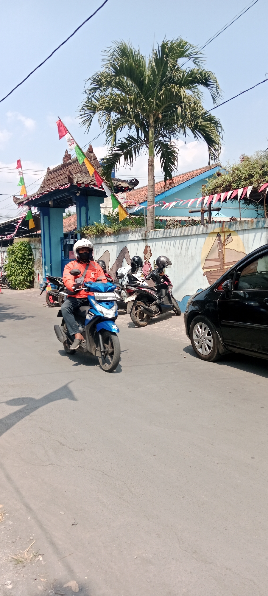 A motorcyclist wearing an orange jacket and a white helmet is riding a blue motorcycle down a paved street. The motorcyclist is facing left, and the motorcycle is moving forward. In the background, there is a blue building with a red-tiled roof, decorated with flags and bunting. A mural is painted on a wall in front of the building. Several other motorcycles are parked in front of the building. A black car is parked on the right side of the street. The scene is set outdoors on a sunny day with a clear blue sky. The location is identified as Purwomartani, Indonesia.