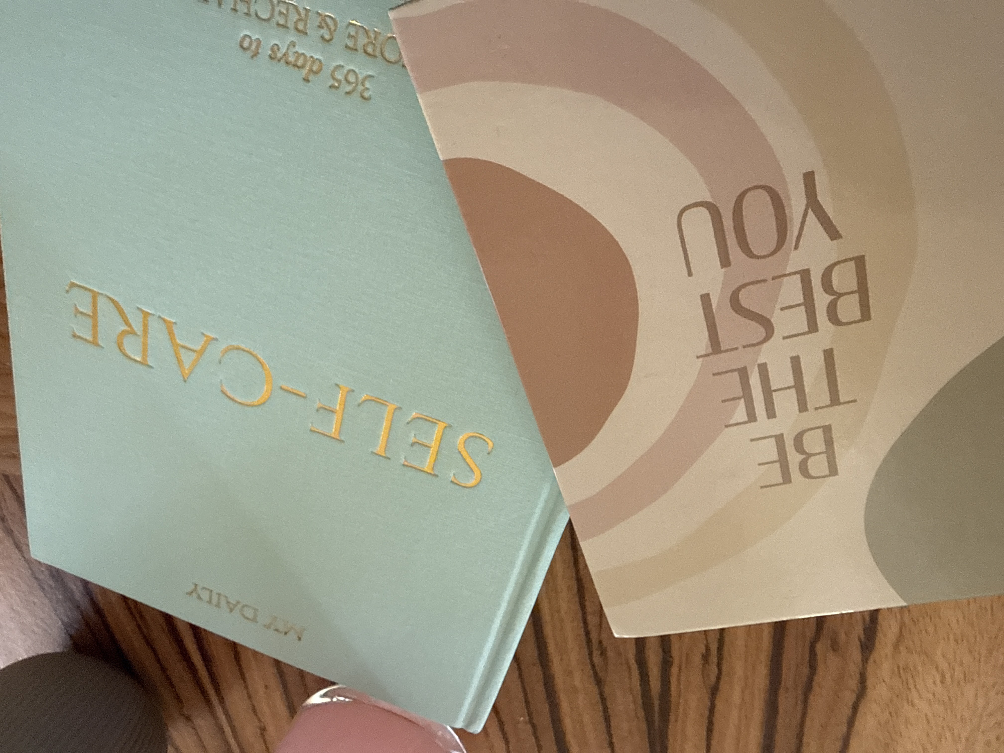 The image shows two notebooks, one blue with the text "SELF-CARE MY DAILY 365 days to ORE & RECH" in gold lettering and the other with a rainbow pattern and the text "BE THE BEST YOU" written in beige lettering. The notebooks are resting on a wooden surface, possibly a table or floor. A fingertip is visible at the bottom of the image. The image does not provide information on the city of Troyan, Bulgaria.