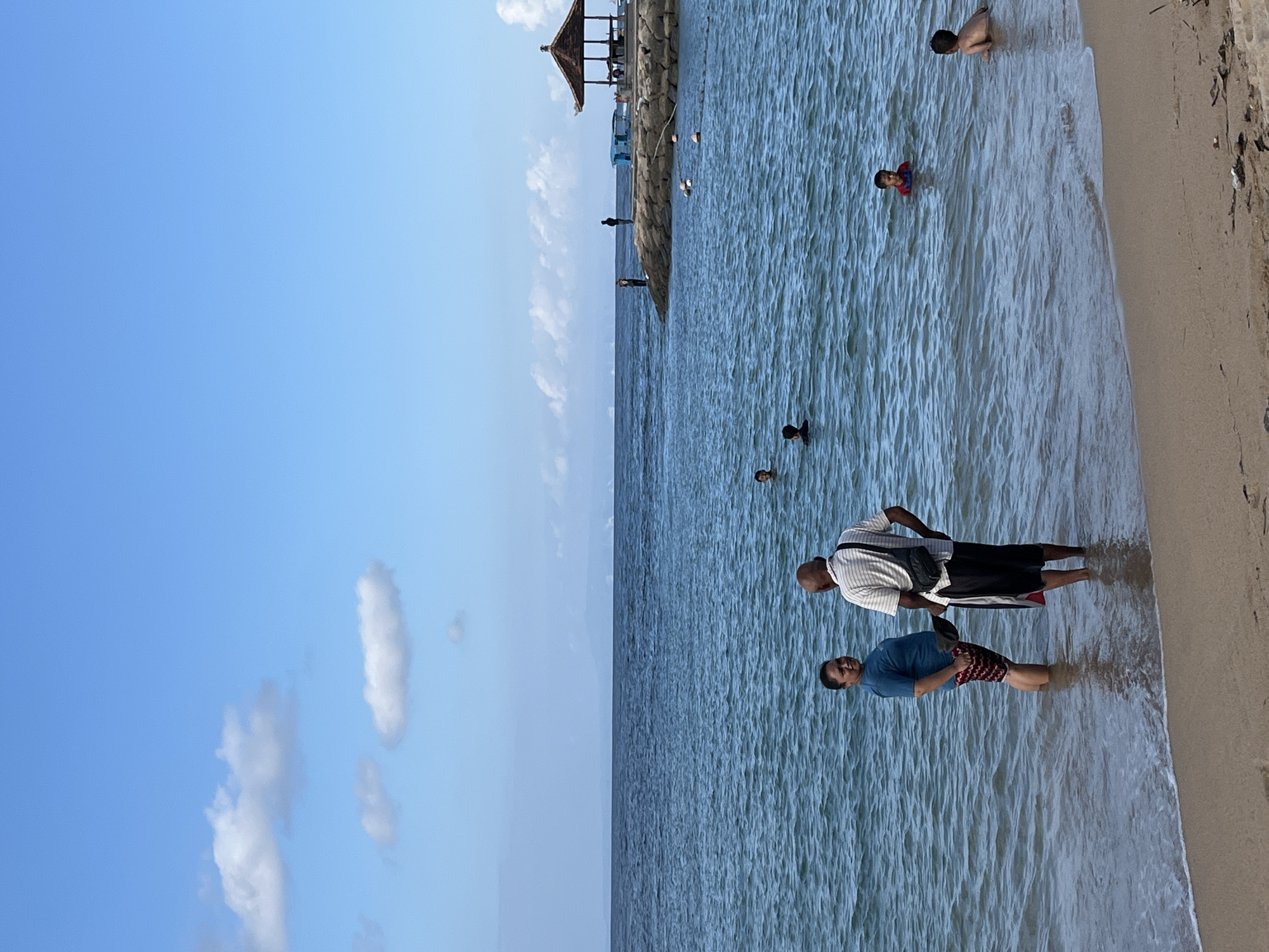 The image depicts a serene beach scene in Kuta Selatan, Indonesia. In the foreground, two adults stand knee-deep in the gentle waves of the ocean. The adult on the left, a woman with dark hair, wears a teal t-shirt and patterned shorts. The adult on the right, a man with a shaved head, is dressed in a striped t-shirt and black shorts, with a bag slung over his shoulder. They are facing each other, engaged in conversation.
Further out in the turquoise water, several children are visible, some swimming and others wading. In the middle distance, a man-made stone pier juts out into the sea, topped with a traditional open-air pavilion with a brown, sloped roof. Two figures stand on the pier, silhouetted against the horizon. The ocean stretches to meet a clear blue sky dotted with a few fluffy white clouds. The beach in the foreground is sandy with some scattered debris. The overall atmosphere is one of peaceful recreation and natural beauty.