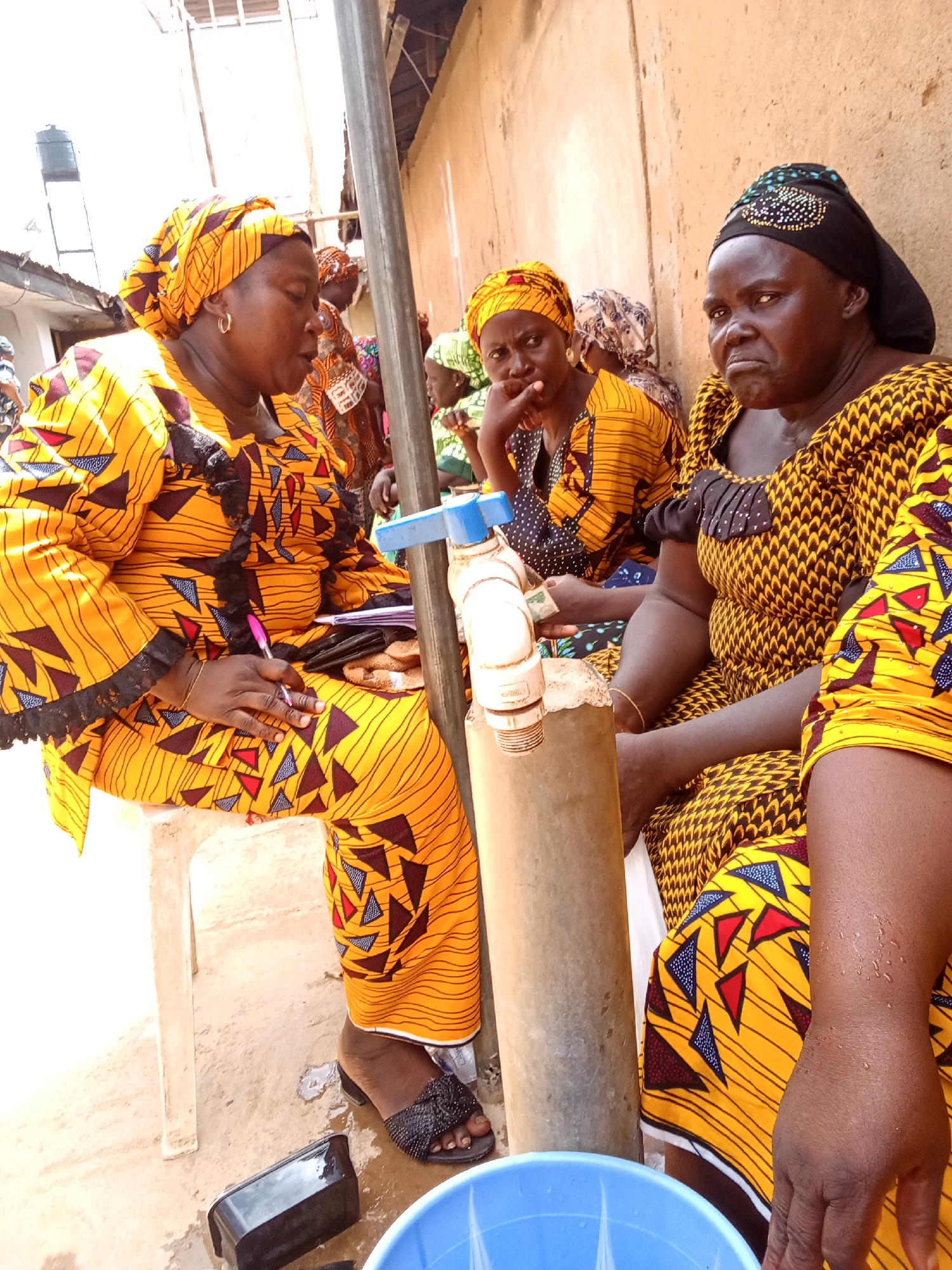 In Sabon Tasha, Nigeria, three women are seated outdoors. The woman on the left, wearing a yellow and orange patterned headscarf and dress, is holding a pen and appears to be writing on a notepad. To her right, another woman in a similar outfit is looking to the left, with her hand near her chin. The woman on the far right, wearing a black headscarf with beaded embellishments and a yellow and black patterned dress, is looking directly at the camera with a stern expression. They are positioned near a large pipe with a blue faucet attachment. In the background, an earthen-colored wall and structures suggest a modest, possibly rural or semi-urban setting. The ground is a mixture of dirt and some wet patches. Several other individuals are visible in the background, also appearing to be seated. The overall scene suggests a community gathering or meeting.