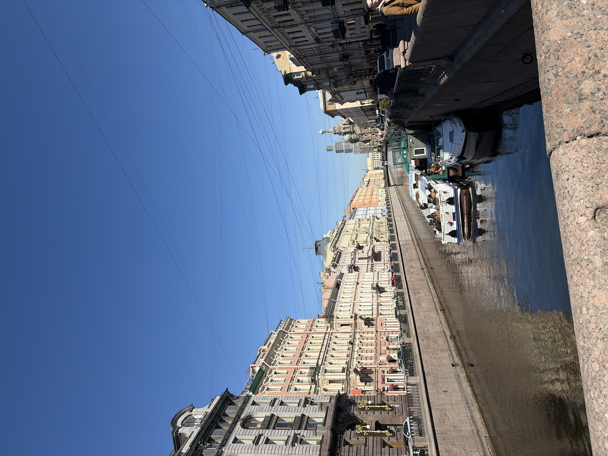 This image depicts a sunny urban canal scene in Saint Petersburg, Russia. The view, taken from an elevated position, looks down a canal lined with historic buildings.
The main subjects include a calm, dark blue canal, several tour boats, and ornate multi-story buildings on both embankments. The canal water reflects the clear blue sky and the surrounding architecture. Multiple tour boats, some with passengers, are visible on the water, indicating active sightseeing.
The buildings lining the canal showcase classic Saint Petersburg architecture, characterized by intricate facades, decorative elements, and varied colors including pastel pinks, creams, and grays. In the distance, a distinctive dome, characteristic of Russian Orthodox churches, rises above the skyline, strongly suggesting the Church of the Savior on Spilled Blood. The canal's edges are constructed with sturdy granite embankments.
The activity taking place centers around tourism, with people enjoying boat tours along the waterway. A few individuals are also partially visible on the embankments.
Notable details include the bright, clear blue sky, indicating excellent weather. The strong shadows cast by the buildings and people suggest it is daytime, likely morning or afternoon. Numerous overhead wires crisscross the sky. The overall scene conveys a pleasant day for exploring the city. No visible text is clearly legible in the image.
