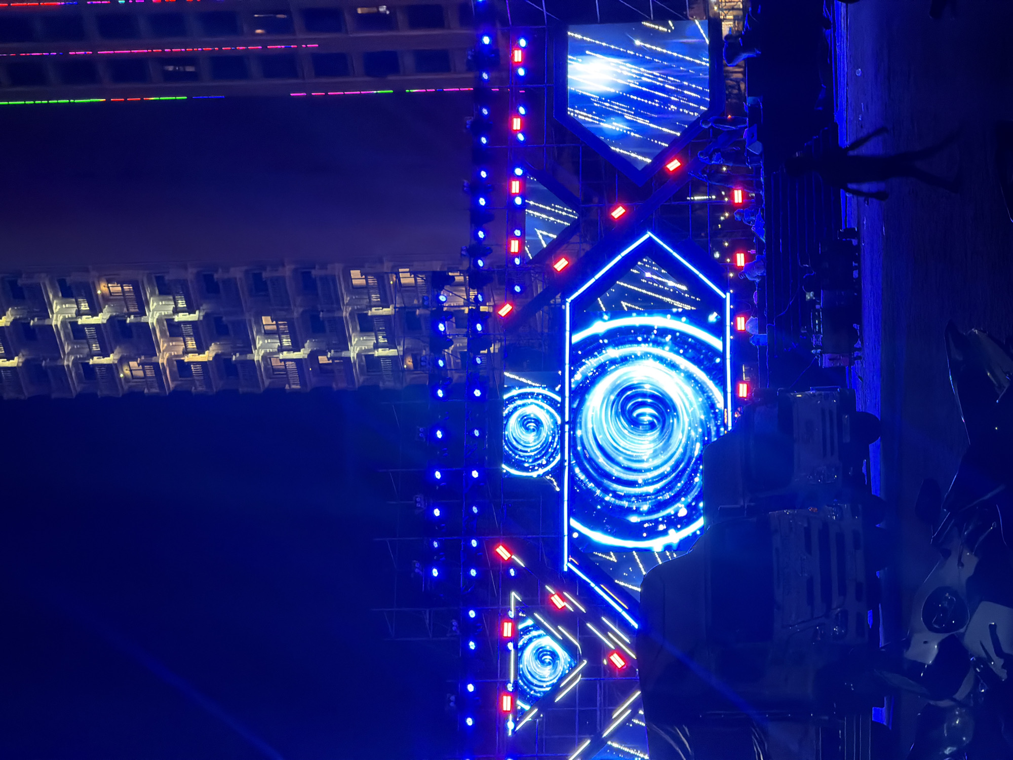 The image depicts a nighttime view of a stage or structure illuminated with blue and red lights. The stage features a series of square or triangular LED panels displaying swirling blue patterns. To the left, a tall building is visible with lit windows. The location is identified as Nha Trang Ward, Vietnam. People are standing on the stage. Equipment is also present, including stage lights. The environment appears to be dark.