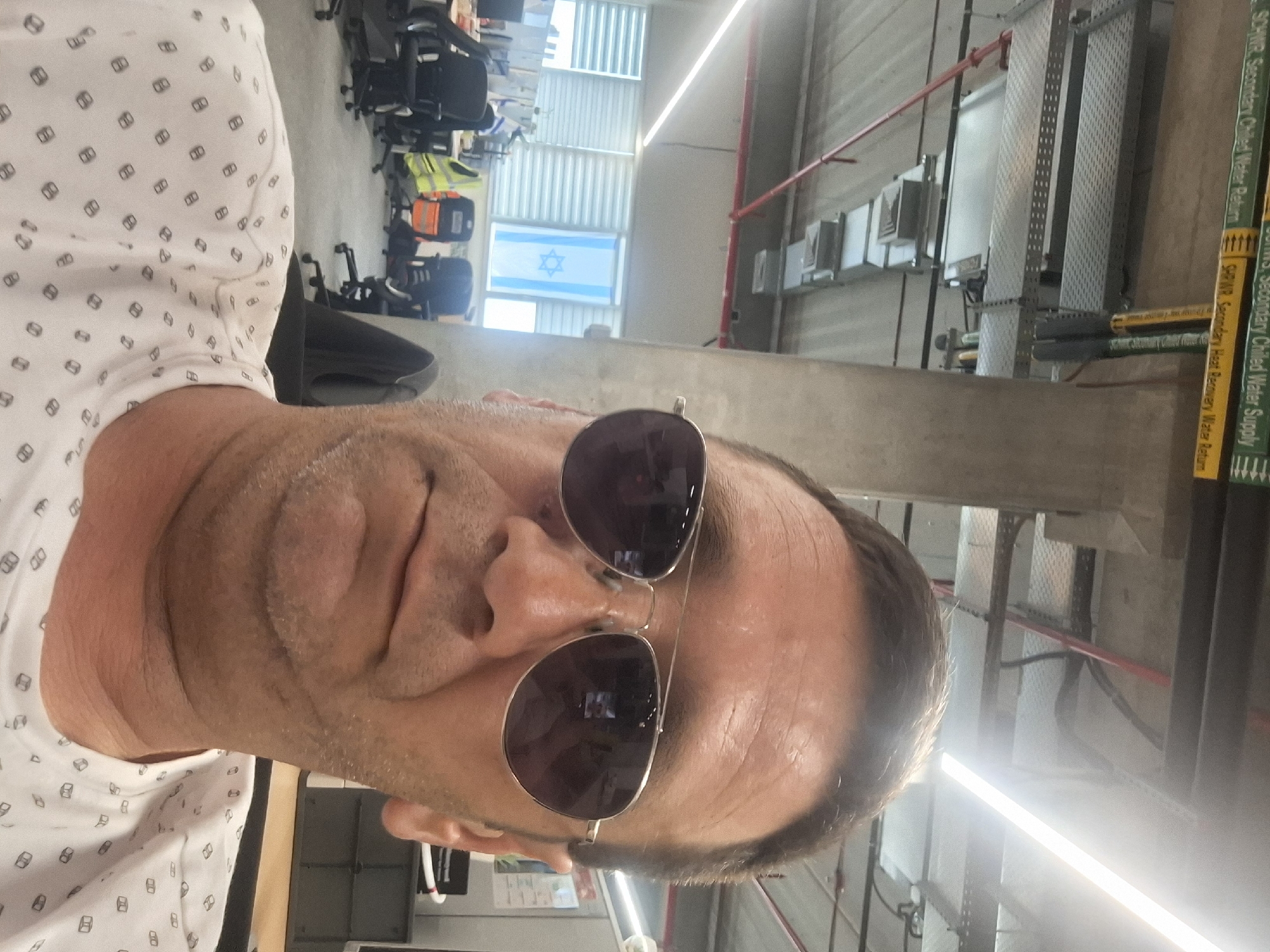 The image shows a man wearing sunglasses and a patterned t-shirt, taking a selfie in what appears to be an office environment. Behind him, an Israeli flag is visible hanging in front of a window with blinds. The office appears to have cubicles with chairs and desks, as well as overhead lighting and conduits. Signs related to "Chilled Water Supply" and "Heat Recovery Water Return" are mounted on the ceiling. The man's face and upper torso are the primary focus of the image.