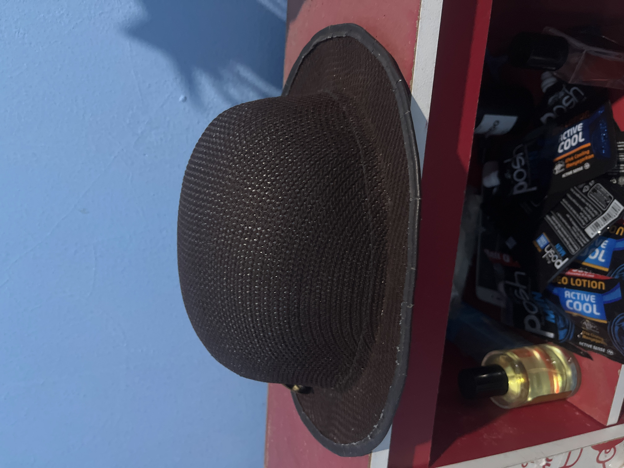 A brown bowler hat with a woven texture rests on a red shelf. The hat has a black brim. Behind the hat, a blue wall is visible. On the shelf below the hat, to the left, is a clear bottle containing yellow liquid and a black cap. To the right of the bottle are several black packages with blue and white text, appearing to be personal care products. The image does not contain any information about the city of Kebumen, Indonesia.
