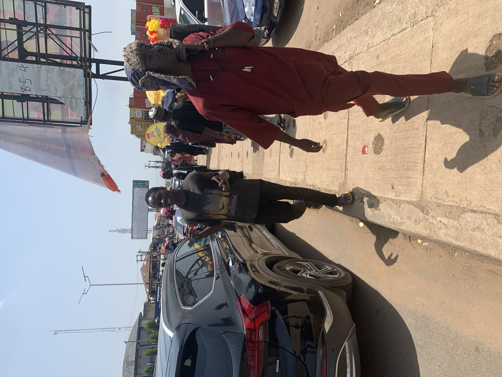 The image captures a street scene in Jos, Nigeria, on a bright, sunny day. In the foreground, two young men are walking on a concrete sidewalk. The man on the left is wearing a dark t-shirt with a graphic print and dark pants, and he is smiling at the camera, with his right hand gesturing. He is also wearing yellow flip-flops. The man on the right is wearing a maroon dashiki and matching pants, and a patterned hat with ear flaps. He has a necklace with black and white beads. He is looking to his right and appears to be talking on his phone, which is held to his ear.
Behind the two men, there is a black SUV parked on the side of the road. The street is lined with various shops and stalls, and there are other people walking in the background. A large billboard with some writing is visible in the upper right portion of the image. The environment appears to be a busy urban area with traffic, buildings, and pedestrians. The overall impression is a snapshot of everyday life in a Nigerian city.