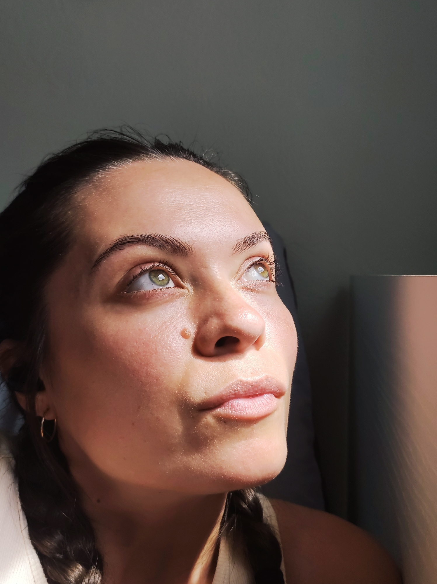 The image is a close-up, head-and-shoulders shot of a woman looking upwards and to her right, illuminated by a shaft of sunlight. Her right eye, with a green iris, is clearly visible, as is a small, raised mole on her cheekbone. Her lips are slightly parted, and her hair is dark and appears to be pulled back, with a braid visible on her right shoulder. She is wearing a simple hoop earring in her visible ear. The background is a muted gray wall, with a portion of what appears to be a lamp or cylindrical object on the right side of the frame, also catching some of the light. The overall impression is one of a candid portrait taken indoors.