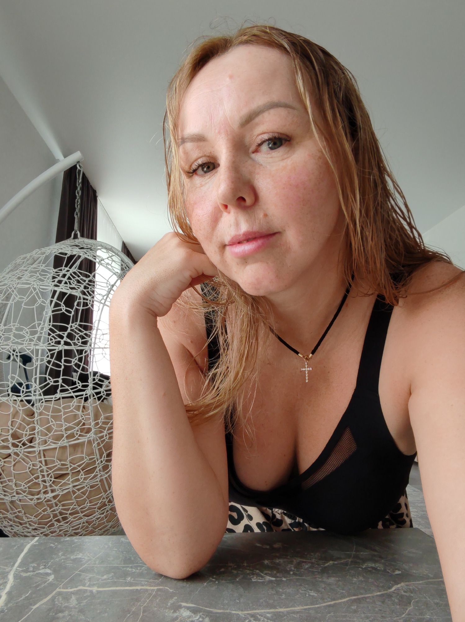 A woman with fair skin and blonde, wet hair is posing for a selfie. She has freckles on her face and is wearing a black top with a cross necklace. Her left arm is bent and resting on a gray marble surface. In the background, there's a white, cage-like hanging chair, and a window with brown curtains. The location is most likely in an apartment in Chaikovsky, Russia, based on the image context.