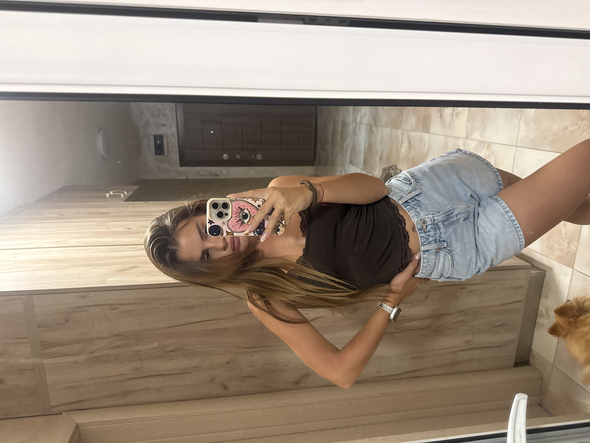 A young woman with long, brown hair is taking a mirror selfie in a room with wooden-paneled walls and tiled flooring. She is wearing a dark brown lace-trimmed camisole and light-wash denim shorts. A smartwatch is visible on her left wrist, and she has a beaded bracelet on her right wrist. Her phone, decorated with a pink case featuring an eye and star design, is held up to capture the reflection. A small, fluffy, light brown dog is visible in the lower left corner of the image, appearing to be moving. The background reflected in the mirror shows a hallway with a closed door and wall-mounted fixtures. The scene appears to be a private residence, likely an apartment or house, in Plovdiv, Bulgaria.