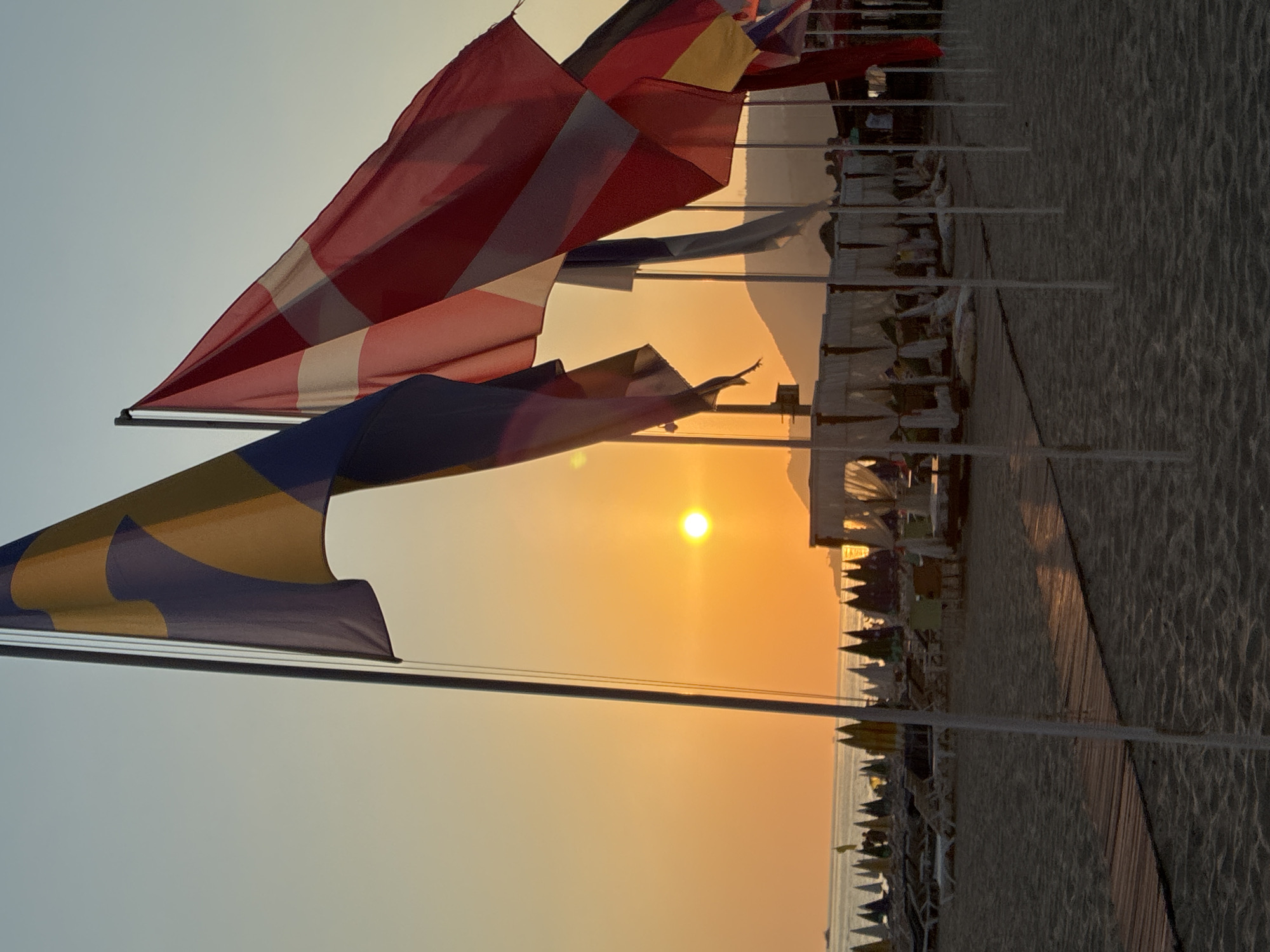 The image captures a serene sunset scene on a beach in Alanya, Turkey. The sky is bathed in warm hues of orange and yellow as the sun dips towards the horizon, casting a soft glow over the water and the sandy shore. Several flags, including what appears to be the flag of Denmark and another with yellow and blue sections, are prominently displayed on flagpoles, their fabric gently swaying in the breeze. In the background, beach umbrellas and lounge chairs are set up, indicating a popular tourist destination. A wooden walkway leads across the sand, and in the distance, hills and mountains form a scenic backdrop. The overall atmosphere is peaceful and relaxing, typical of an evening at a coastal resort.