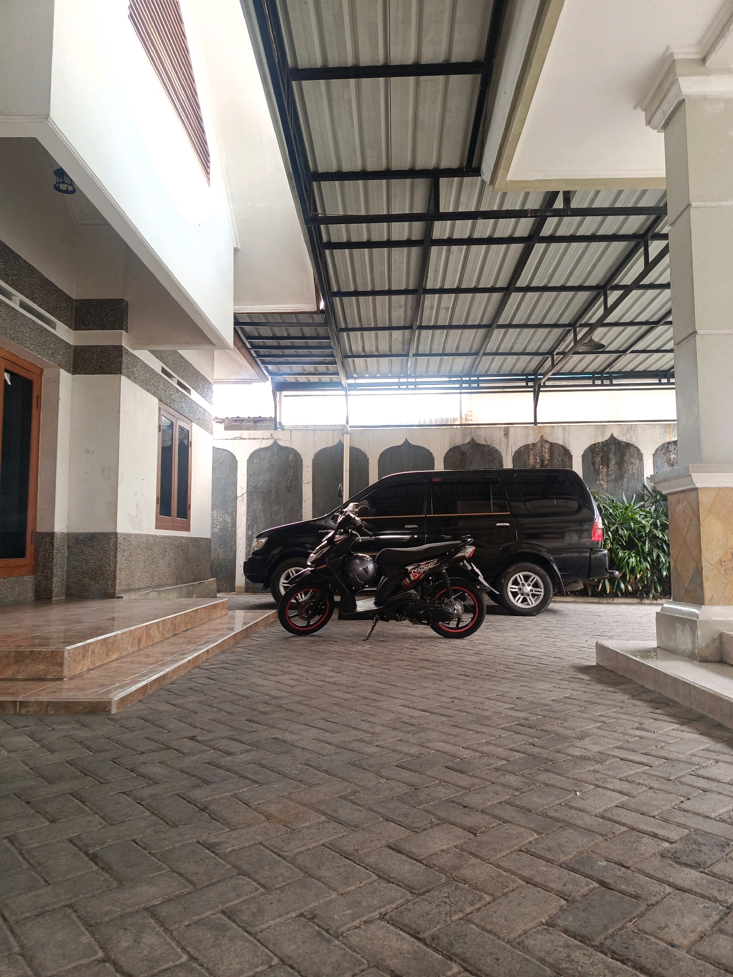 This image depicts the outdoor area of a building in Kebonagung, Indonesia, likely a residential or commercial property. The foreground is paved with interlocking grey brick pavers laid in a herringbone pattern. To the left, a multi-story building with a white facade and a textured brown lower section features a wooden-framed window and a set of tiled stairs leading up to an entrance. A white overhang with decorative molding is visible above the stairs.
Dominating the mid-ground is a black SUV parked on the brick paving. A black scooter with red accents is parked in front of the SUV. Behind the vehicles, a textured grey wall with arched decorative elements runs horizontally. To the right of the wall, lush green foliage is visible.
The background features a corrugated metal roof supported by a black metal frame, suggesting a covered parking area or outdoor living space. The overall impression is of a functional and maintained property in a tropical or subtropical climate, given the presence of the greenery and the style of the building. There are no people or animals visible in the image.