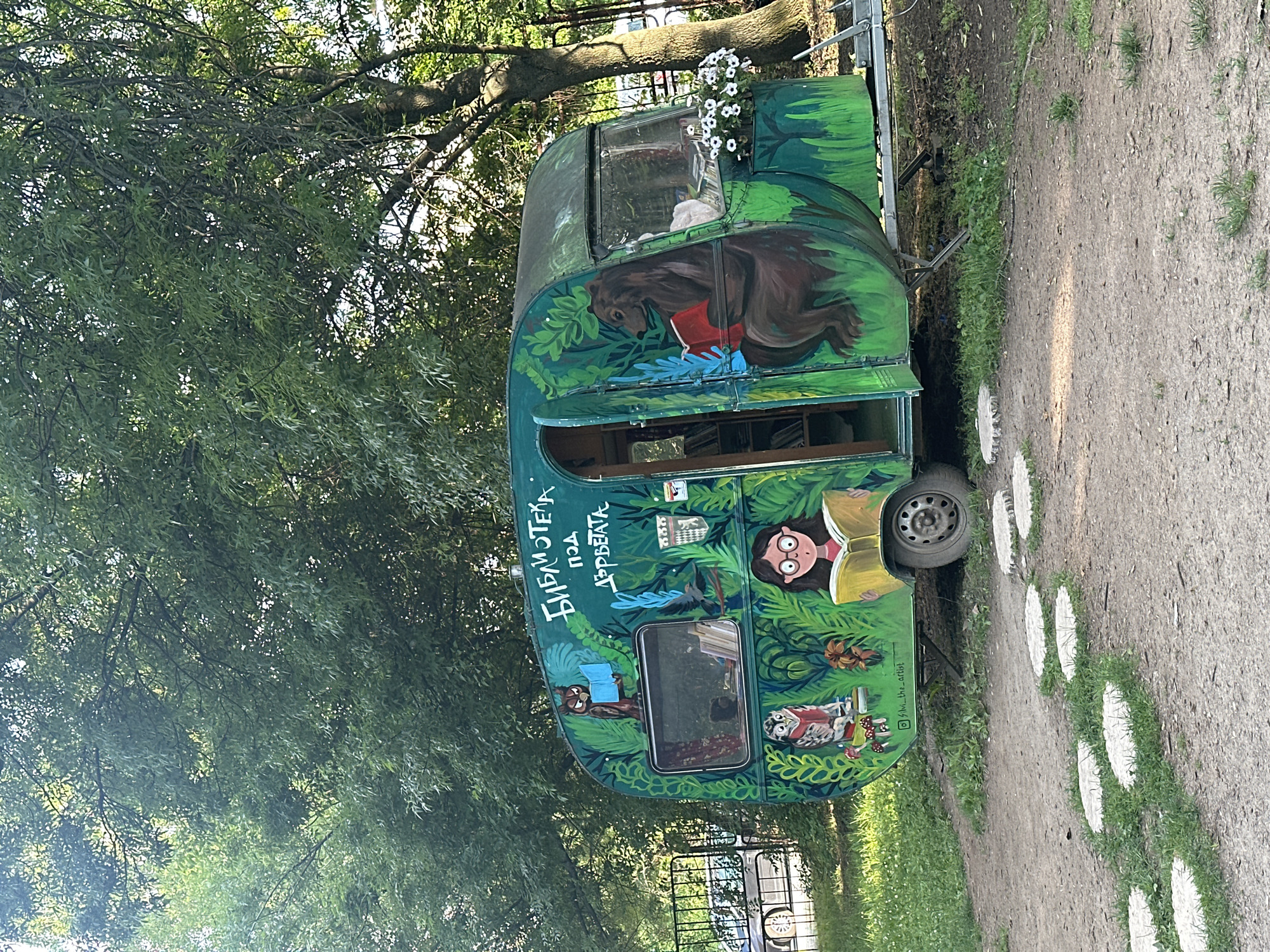 A vibrant, green-painted caravan, transformed into a mobile library named "Библиотека под дърветата" (Library under the trees), is parked on a dirt path with grass and stepping stones leading to it. The caravan features whimsical painted murals of animals reading books, including an owl, a girl with glasses, and a bear. One side of the caravan is open, revealing shelves stocked with books inside. White flowers in a planter adorn the front corner of the caravan. The caravan is surrounded by lush green trees and foliage, suggesting an outdoor setting. In the background, a metal fence and a car are visible. This scene is located in Sofia, Bulgaria.