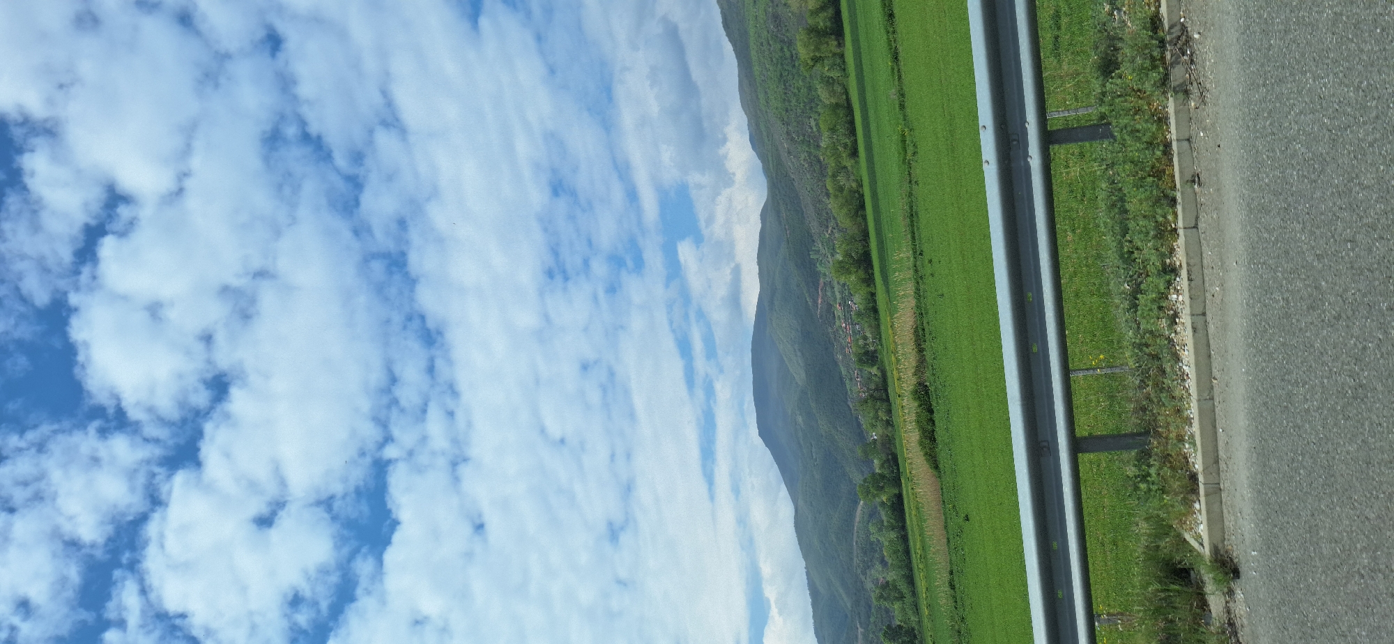 The image depicts a roadside scene, likely taken from a moving vehicle. On the left, a vibrant blue sky is filled with numerous white, puffy clouds. Adjacent to the sky is a mountain covered with trees and vegetation. A large green field, possibly farmland or a meadow, extends towards the right. A metal guardrail runs alongside the field, separating it from the road. A grey asphalt road occupies the rightmost section of the image, with a narrow strip of grass and gravel between the guardrail and the road. There are no people visible in the scene. The location could be Valkovo, Bulgaria, considering the landscape features and vegetation typically found in this region.