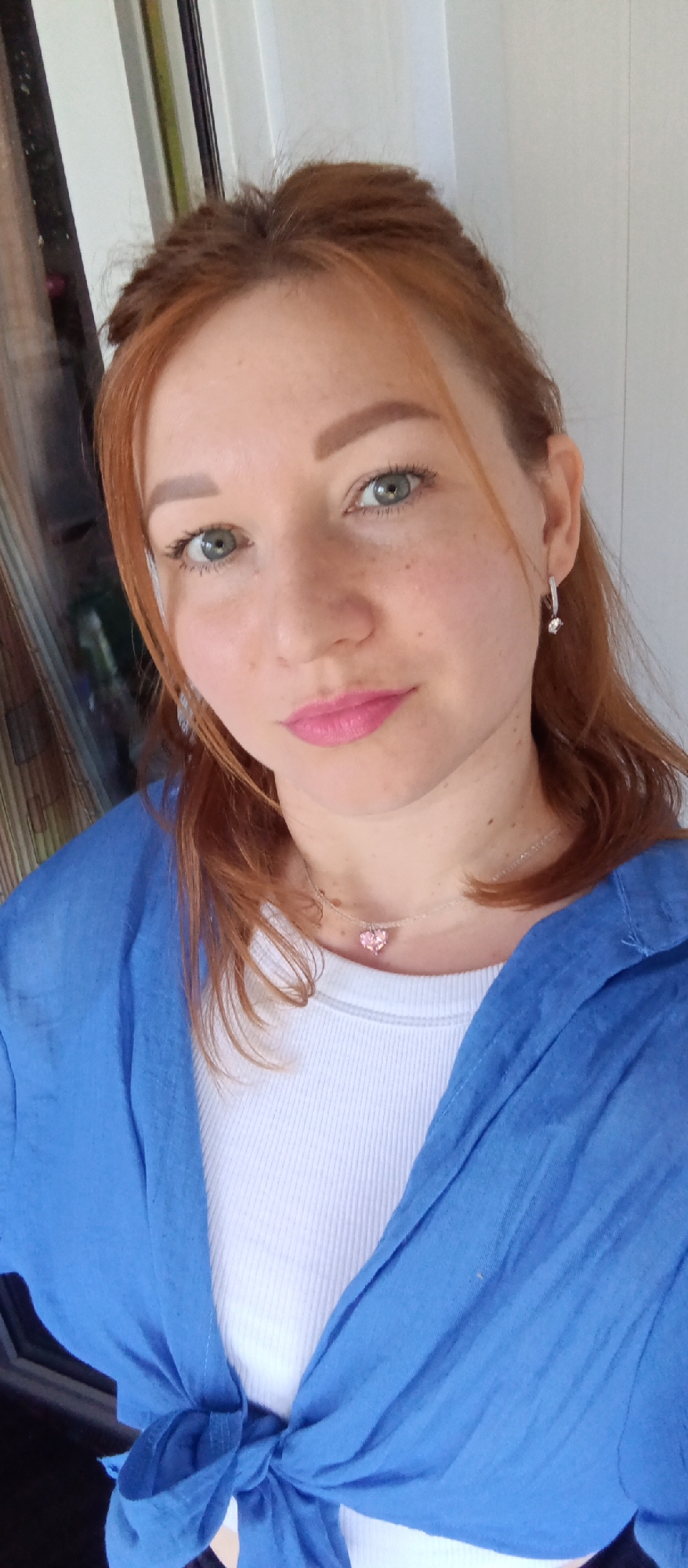 A woman with auburn hair, wearing a white ribbed tank top and a bright blue button-down shirt tied at the waist, is centered in a selfie. She has fair skin with freckles and is wearing pink lipstick. Her green eyes look directly at the camera. She also wears silver drop earrings and a delicate silver necklace with a pink heart pendant.
Behind her, a blurred window with vertical dark bars and a white curtain can be seen, suggesting an indoor setting, possibly in the city of Unknown, Unknown. The lighting is soft and natural, illuminating her face and the details of her clothing and accessories.