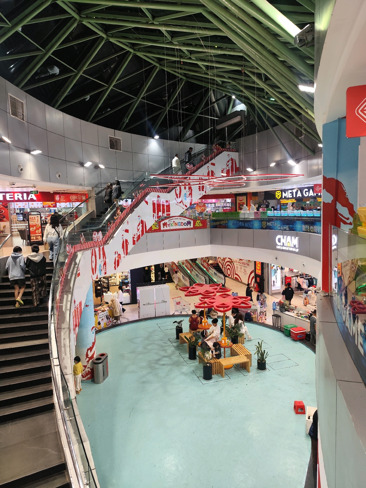 An interior view of a multi-level shopping mall in Da Lat, Vietnam, shows a wide atrium with a light blue floor. On the left, a wide staircase ascends, with several shoppers walking up. The staircase is decorated with red and white striped banners featuring cartoonish figures. On the right, a section of the mall's facade is visible, with a large red sign. In the center of the atrium, several groups of people are gathered around low wooden tables and benches, some seated and some standing. Red, flower-shaped umbrellas provide shade. Various shops are visible on the upper levels, with signage including "META GAME" and "CHAM CAFE & NEST." Escalators are also visible on the lower levels. The ceiling is a complex network of green metal beams and lights. A child in a yellow outfit stands near a silver trash can by the staircase.
