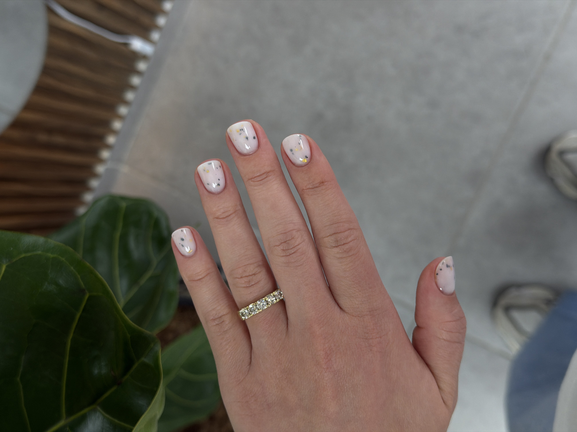 A woman's hand is prominently featured, adorned with a gold ring set with multiple stones. The nails are painted with a light, speckled design. The background includes a plant with broad, green leaves on the left and a blurred gray surface with a hint of a shoe in the upper right. The location is not discernible from the image.