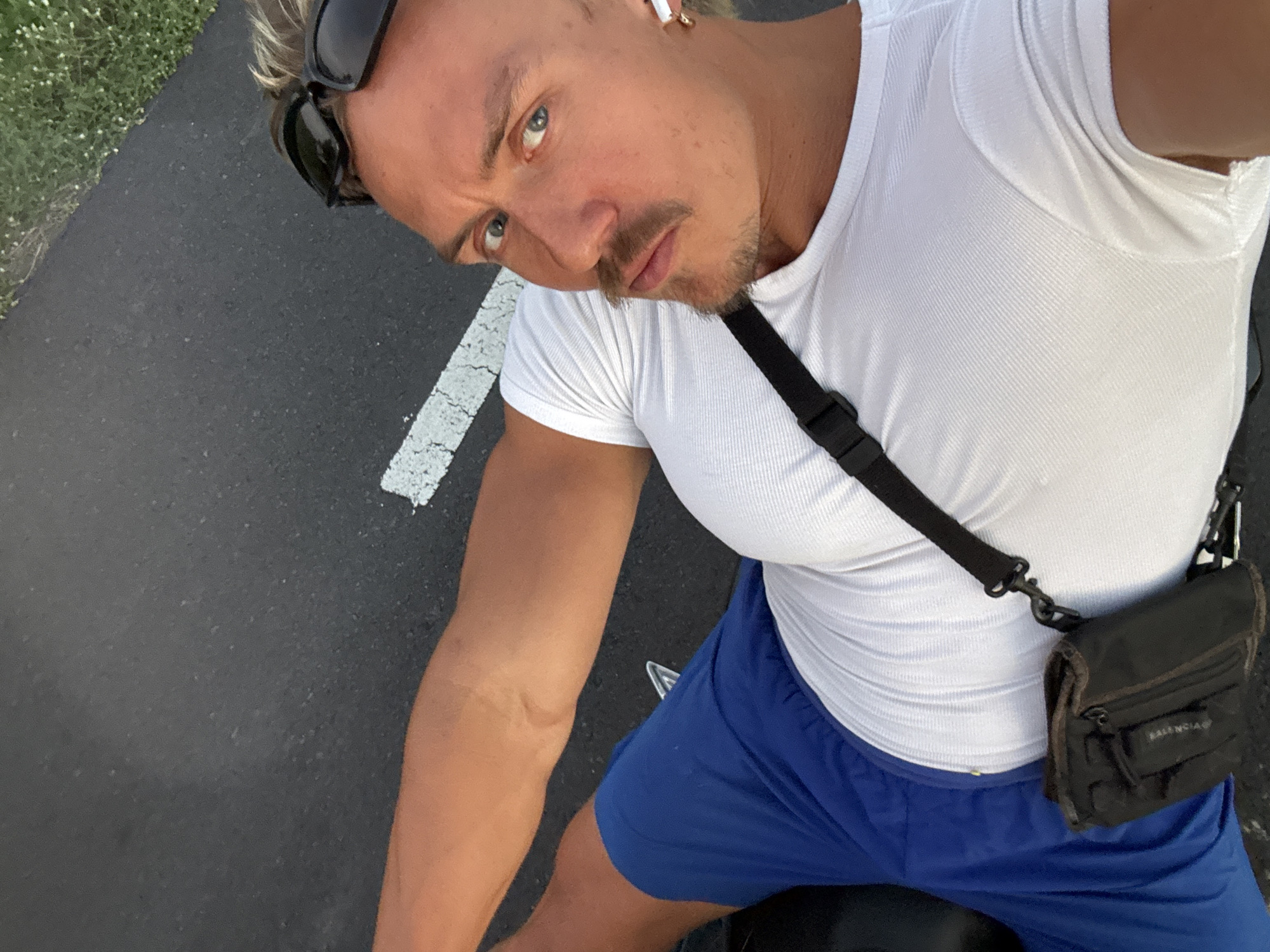 A man with blonde hair and blue eyes, wearing a white ribbed tank top and blue athletic shorts, poses outdoors on asphalt with a white stripe, seemingly in daylight. Sunglasses are perched on his head. He wears an earpiece in one ear and a black crossbody bag. The location is consistent with an outdoor road or path.