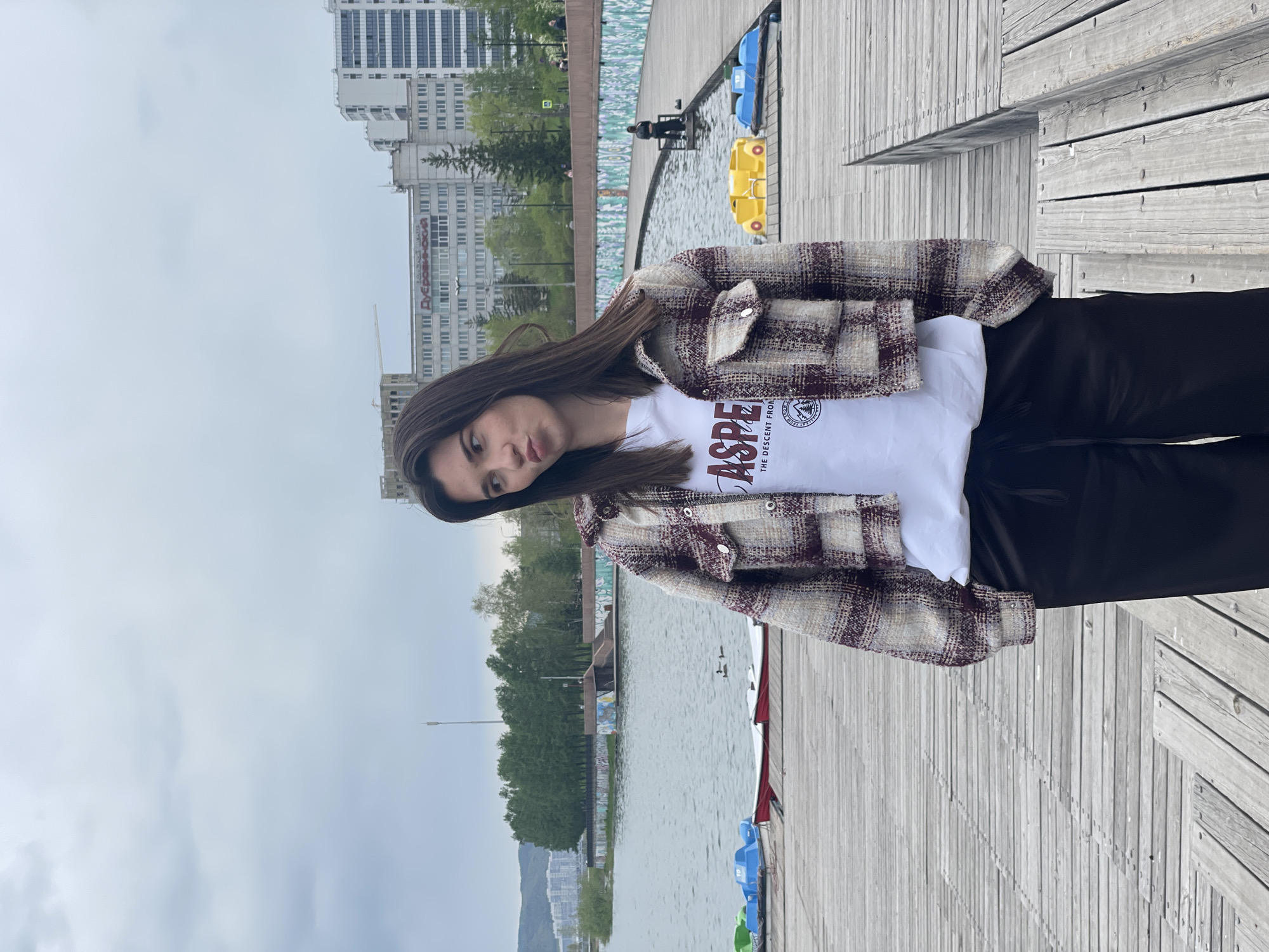 A woman is standing on a wooden boardwalk adjacent to a body of water. The woman is wearing a white t-shirt, a plaid jacket, and dark pants. In the background are buildings, trees, and the sky. The location is in Krasnoyarsk, Russia. Paddle boats are visible in the water.