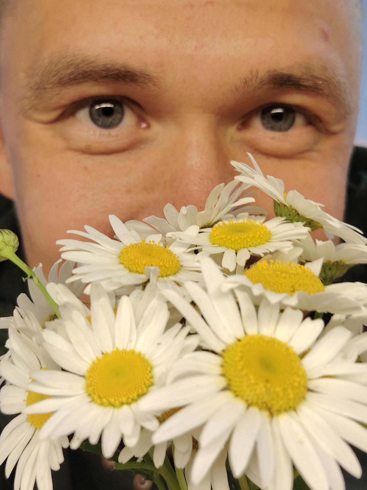 A close-up shot focuses on a person's eyes and a bouquet of daisies. The person, viewed from the chest up, has light-colored eyes and fair skin with a scattering of freckles. Their eyebrows are visible, as are the subtle lines around their eyes, suggesting a gentle expression. The upper portion of their head is cut off by the frame.
In front of the person's face, a vibrant bouquet of white daisies with bright yellow centers is prominently displayed. The flowers are in various stages of bloom, with some fully open and others still budding. The focus shifts between the person's eyes and the flowers, creating a sense of depth.
The lighting suggests an outdoor setting, possibly during the day, with soft illumination. There are no explicit location cues in the image to suggest Chaikovsky, Russia, or any specific weather conditions. The overall mood is gentle and natural. No text is visible in the media file.