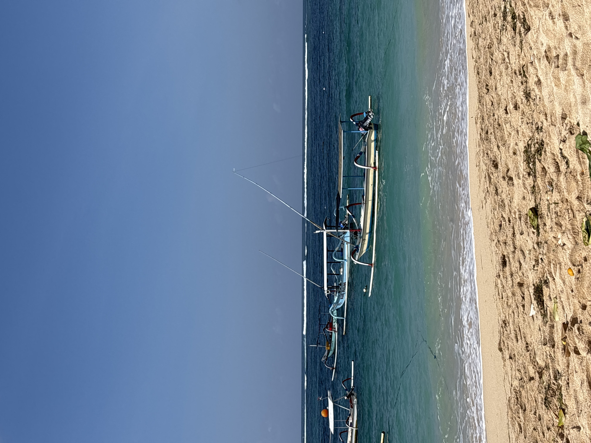 A sunny day at the beach in Kuta Selatan, Indonesia, features calm, turquoise waters lapping against a sandy shore. Several traditional Balinese fishing boats, known as jukung, are anchored in the shallow water. These boats are characterized by their outriggers, which provide stability, and many have canopies for shade. One boat in the foreground is light yellow with blue trim and a small outboard motor. Further out, a light blue jukung with the words "BALI JELOG" visible on its side is also present. The sky above is clear and blue, with a distinct horizon line where the water meets the sky, marked by a few faint white waves in the distance. The sandy beach in the foreground is textured with ripples and scattered with dry seaweed and a few small pieces of debris. No people are visible in the frame, indicating a peaceful, unoccupied scene.