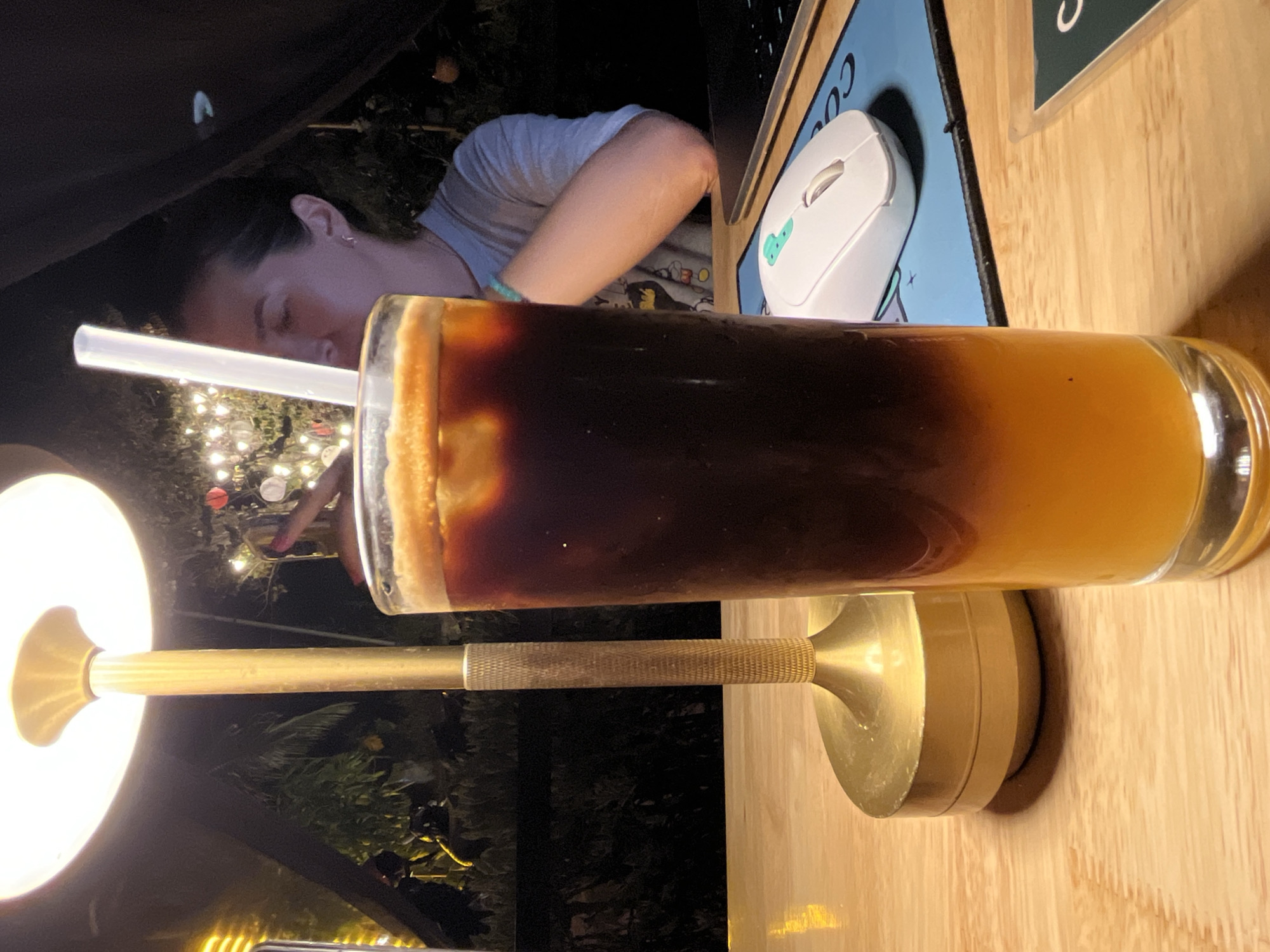 A person is sitting at a table with a drink in a glass, likely an iced coffee, containing a straw. A computer mouse is on the table, sitting on a blue mousepad. The drink is illuminated by a lamp. The photo was likely taken outdoors in Bac Nha Trang Ward, Vietnam.