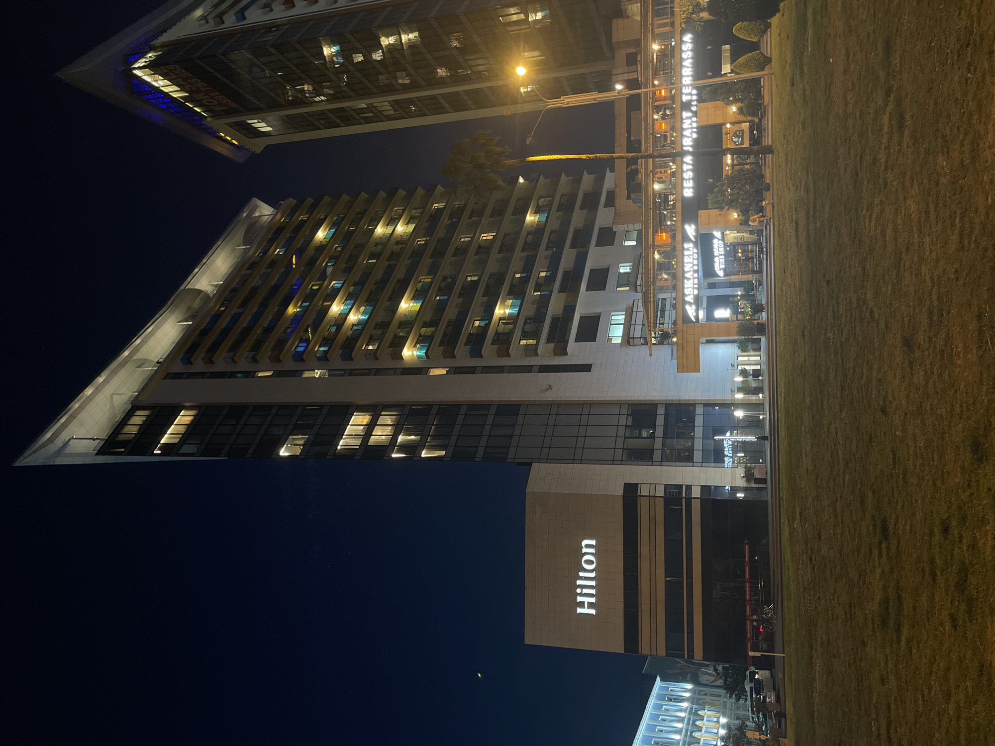 The image shows a nighttime view in Batumi, Georgia, featuring the Hilton hotel. The hotel's sign is visible on the building. Nearby is a restaurant, "RESTAURANT TERRASSA." A grassy area stretches to the right of the buildings. Streetlights illuminate the scene. Other multi-story buildings are visible in the background.